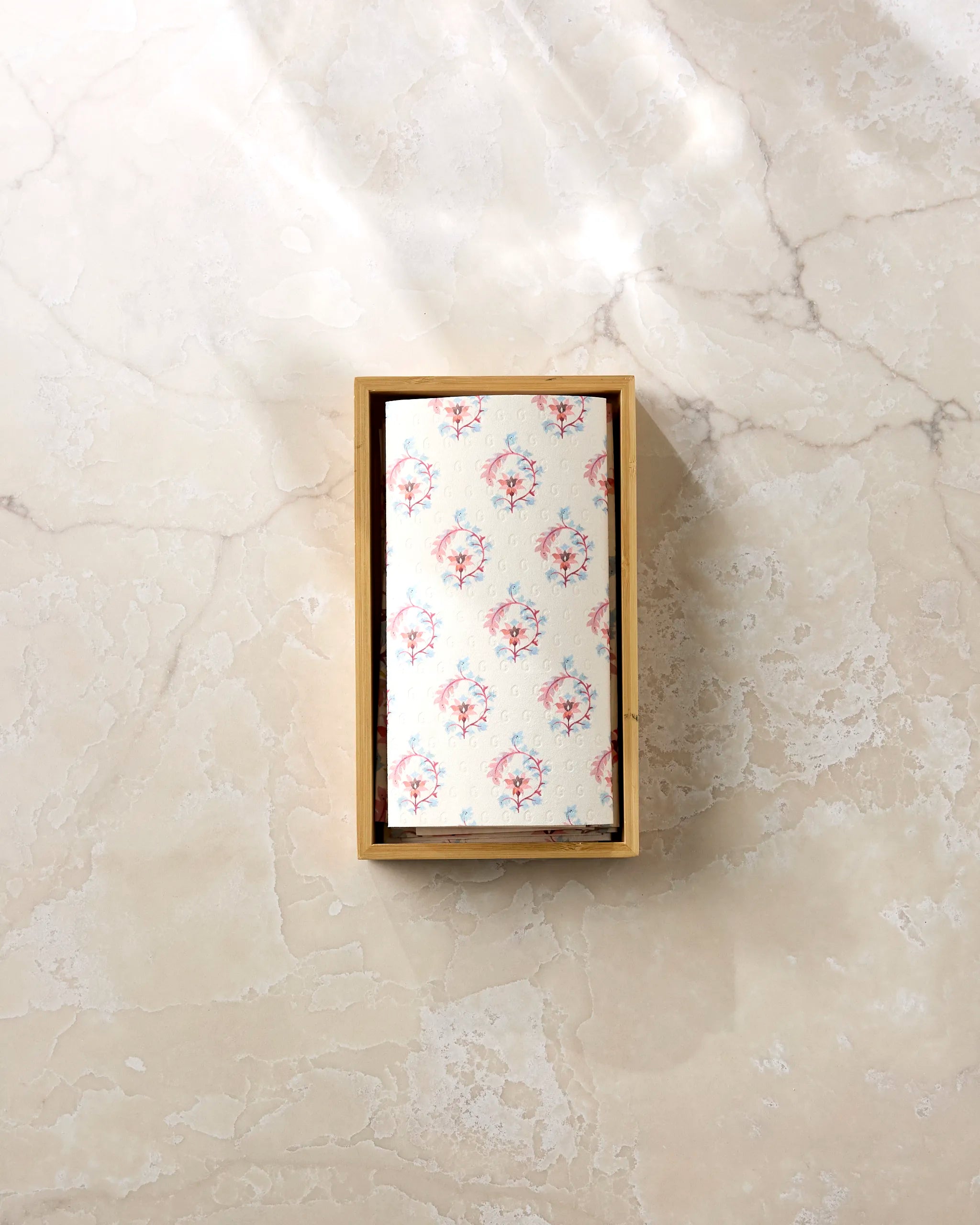 The Folk Medallion by Maayan Alper-Swan is a rectangular wooden box with a delicate red and blue floral design, perfect for stylishly presenting eco-friendly paper towels on any light beige marble surface.