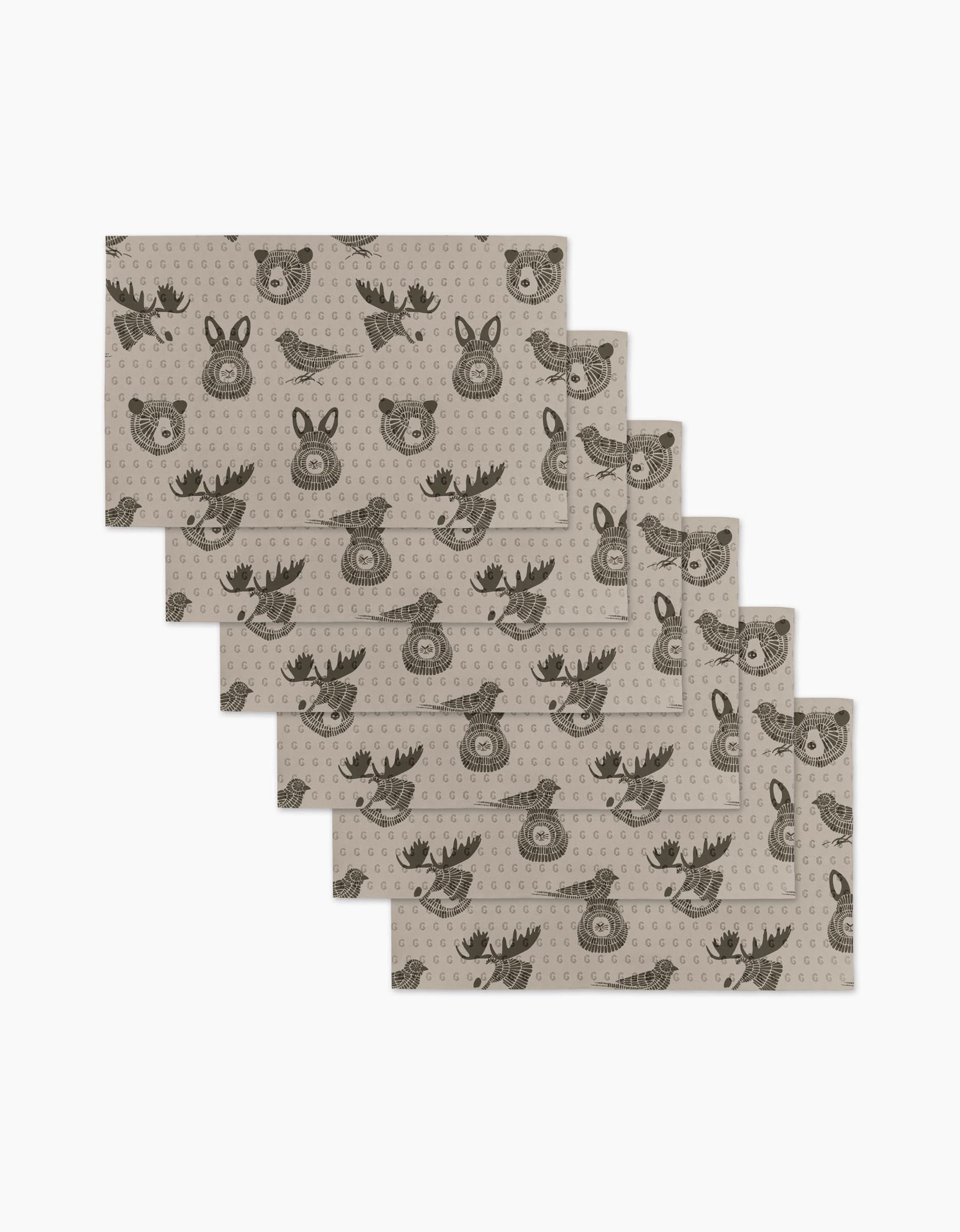 Six beige Forest Animals placemats by Rosa Kichline feature repeating cartoon faces of a bear, deer, and fox in dark brown. These eco-friendly kitchen towels are arranged in a slightly fanned-out stack.