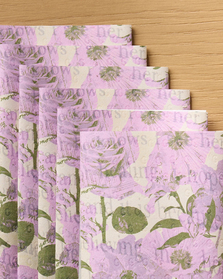 Geometry’s Forever Mom eco-friendly kitchen towels, featuring pastel floral and text prints in lavender, green, and beige, are folded and fanned out on a wooden surface.