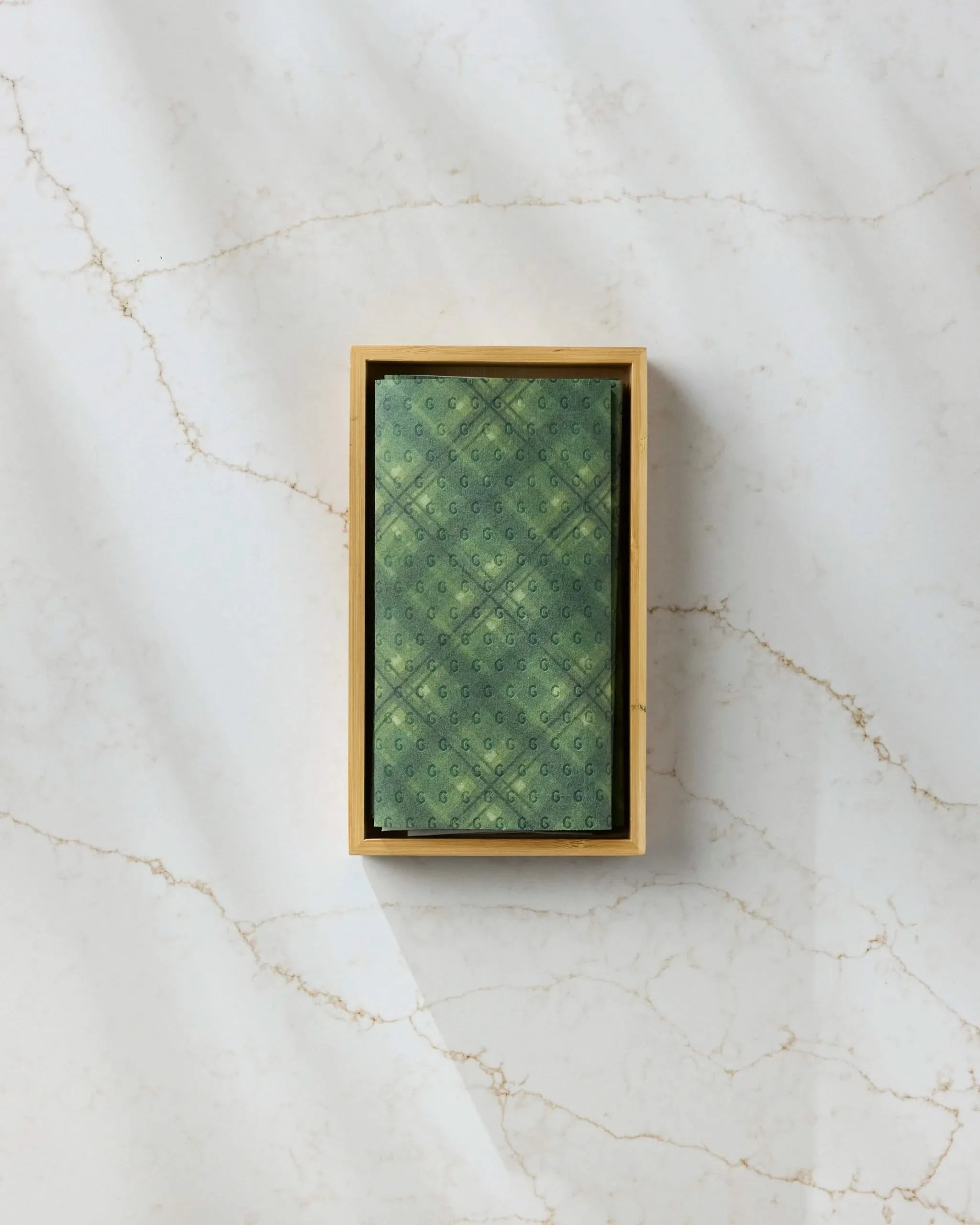 A Geometry Forever Plaid reusable towel with a green pattern rests inside a wooden box on a white marble surface featuring light brown veining.