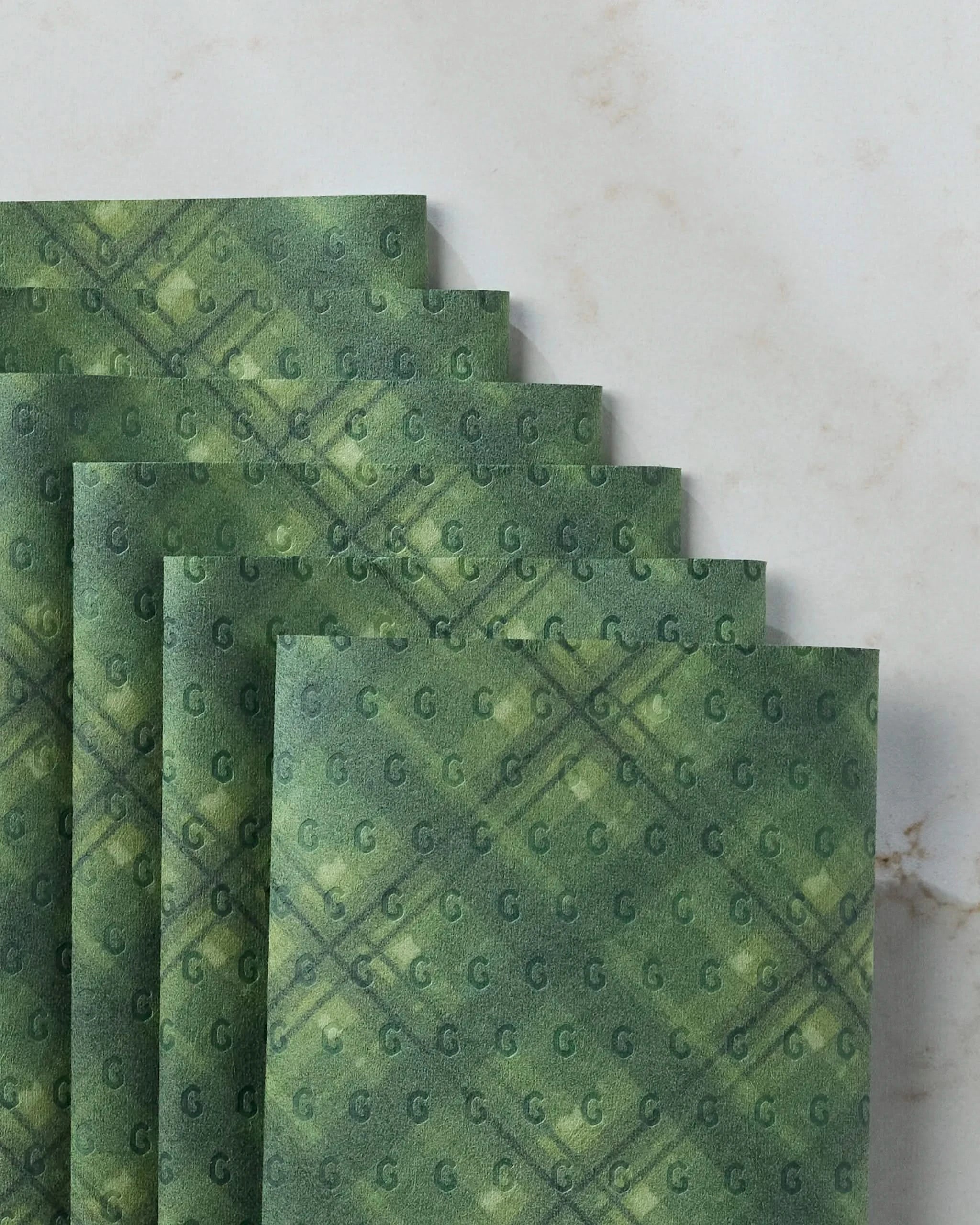Five green Forever Plaid napkins by Geometry, featuring a plaid pattern with small G letters, are styled in a fan shape on white marble. These eco-friendly, reusable napkins add charm to any table setting.
