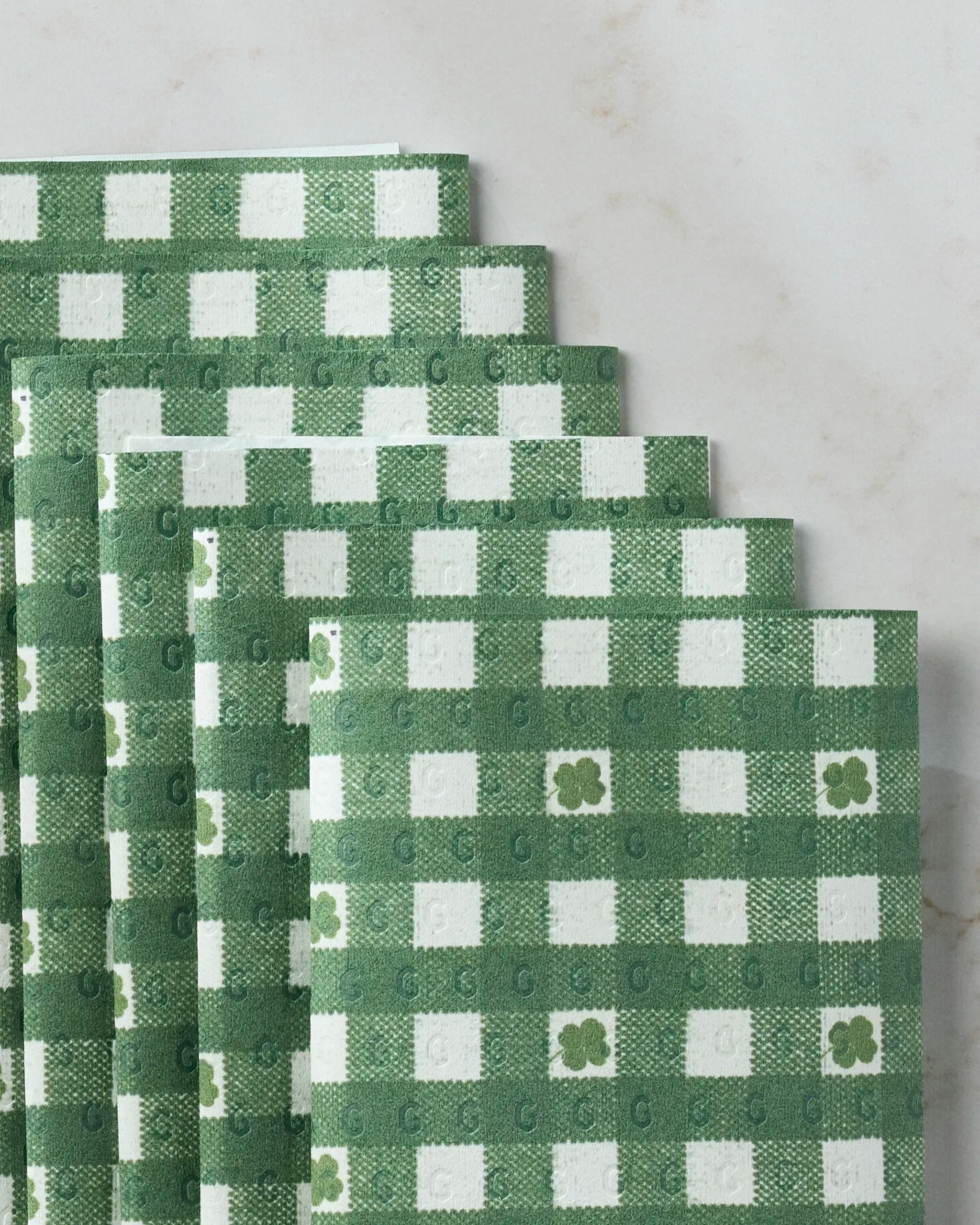 Geometry’s Fortunate Gingham reusable paper towels, featuring a green and white checkered pattern with small flowers and “G” monograms, are fanned out on a light marble surface.
