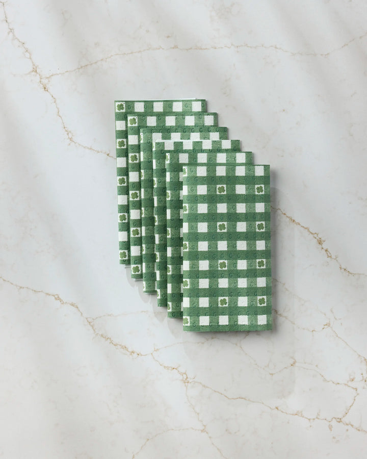 Five Fortunate Gingham reusable towels by Geometry, featuring green and white checkered patterns, are neatly stacked on a white marble surface with light brown veining.