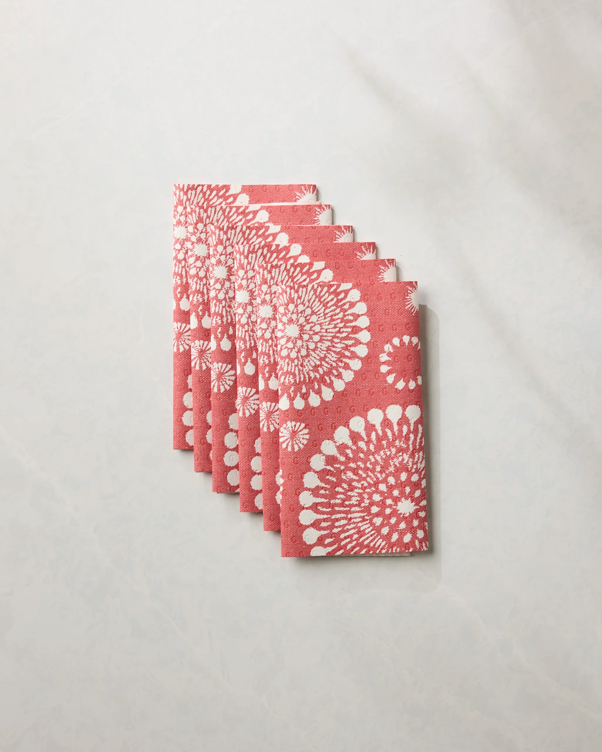 Six neatly folded Freedom Sparks Bright reusable paper towels by Geometry, featuring a coral tone with a white floral burst pattern, are stacked on a light, marble-like surface.