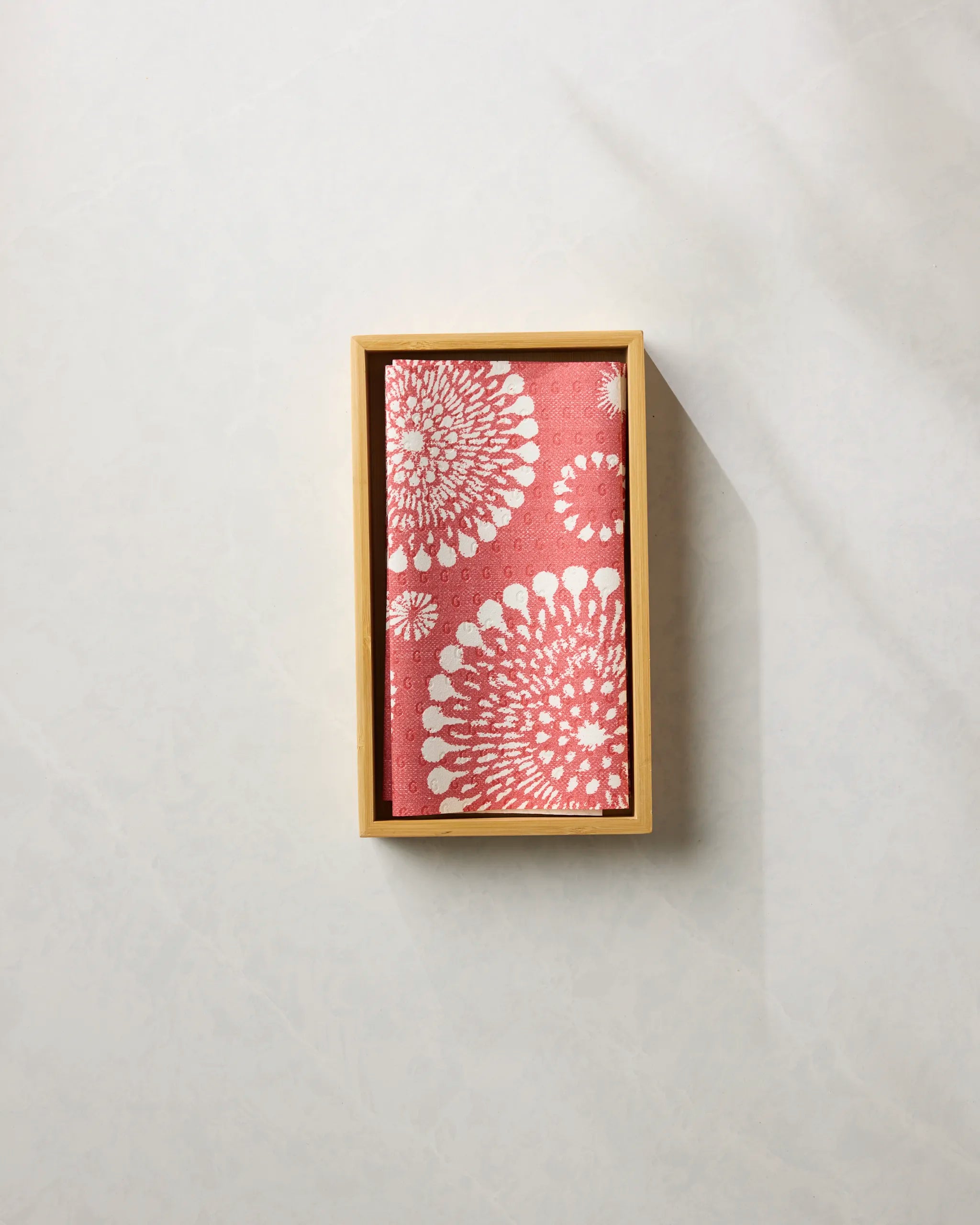 The Freedom Sparks Bright by Geometry, a pink cloth with white floral patterns resembling eco-friendly reusable paper towels, is neatly placed in a rectangular wooden box on a light, textured surface.