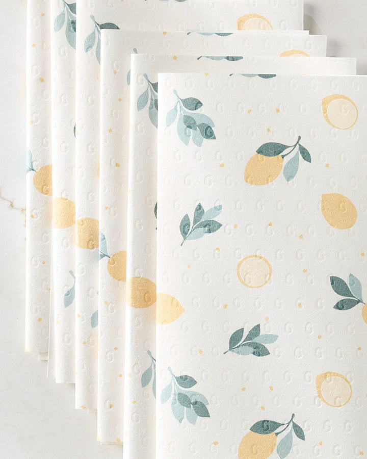Several sheets of Sara Mari’s Freshly Picked eco-friendly kitchen towels, featuring a yellow lemon and green leaf pattern, are fanned out on a light-colored surface.