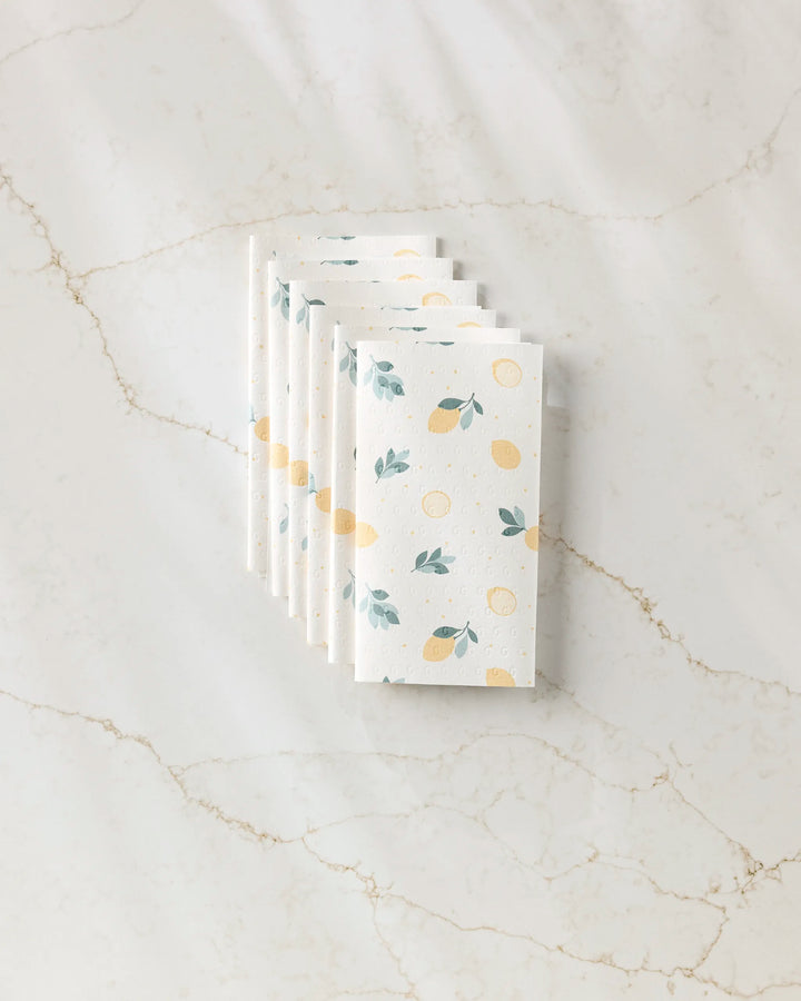 Seven folded, reusable "Freshly Picked" paper towels by Sara Mari, featuring a lemon and leaf pattern, are stacked neatly on a white marble surface with subtle beige veining.