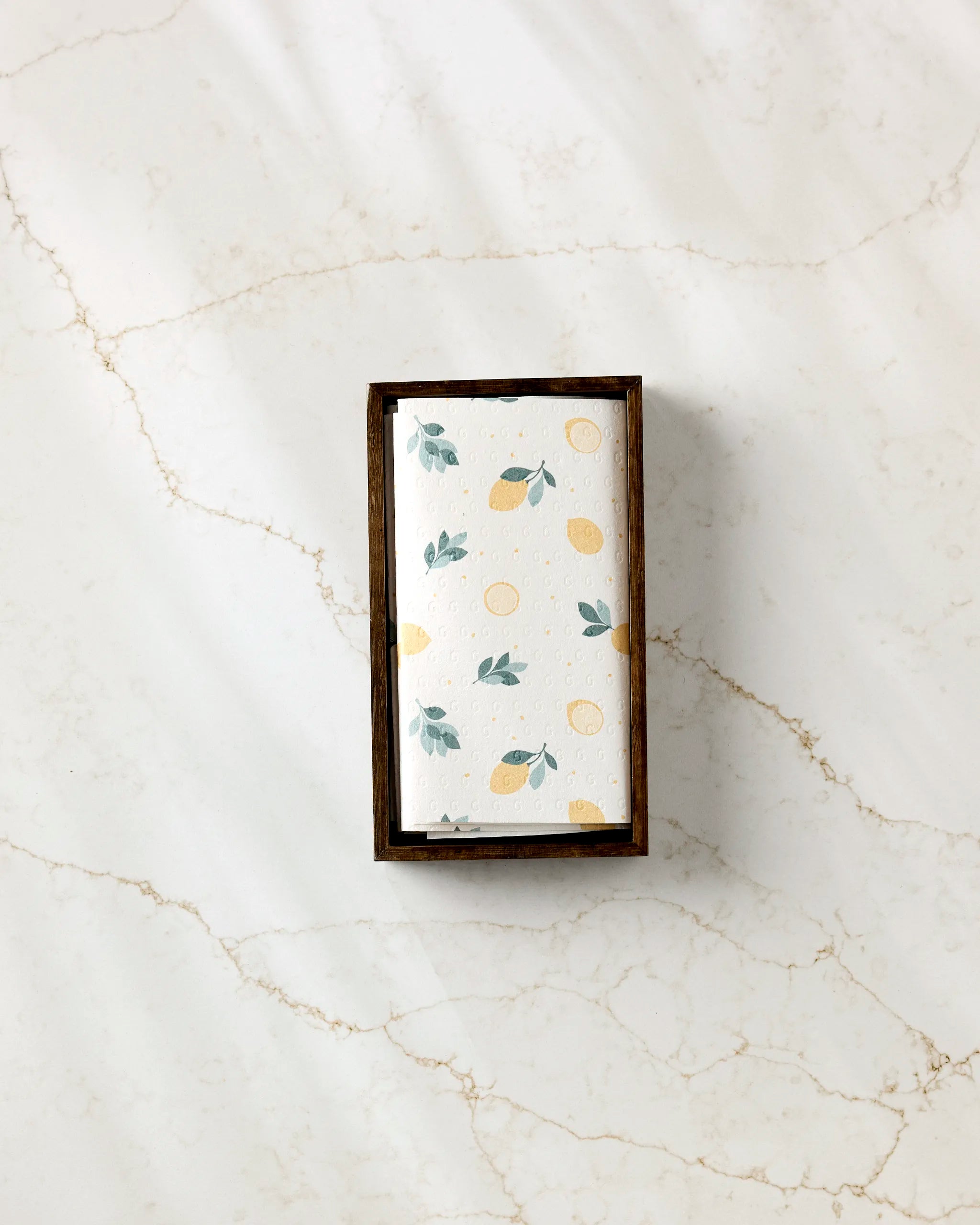 The Sara Mari Freshly Picked bar soap, wrapped in white with yellow lemons and green leaves, sits in a wooden tray on white marble with tan veins next to eco-friendly kitchen towels.