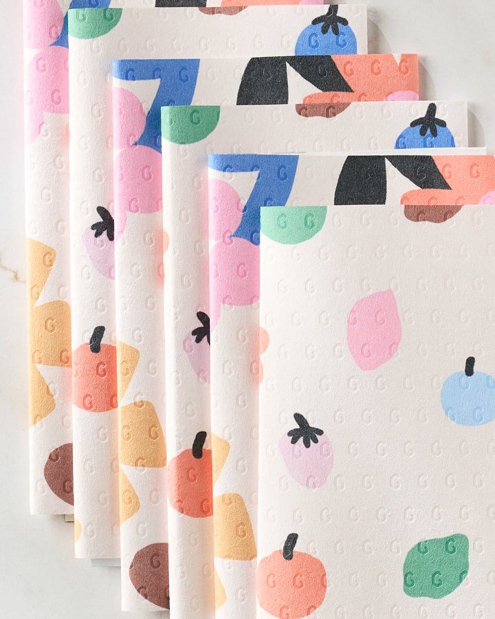 Five eco-friendly Fruit Basket paper towels by Jennifer Bouron, each featuring colorful fruit illustrations and a subtle embossed G pattern, are fanned out on a light surface.
