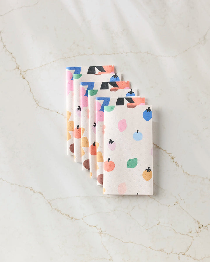Six ‘Fruit Basket’ rectangular sponges by Jennifer Bouron, each featuring colorful abstract fruit designs, are lined up diagonally on a light marble surface.