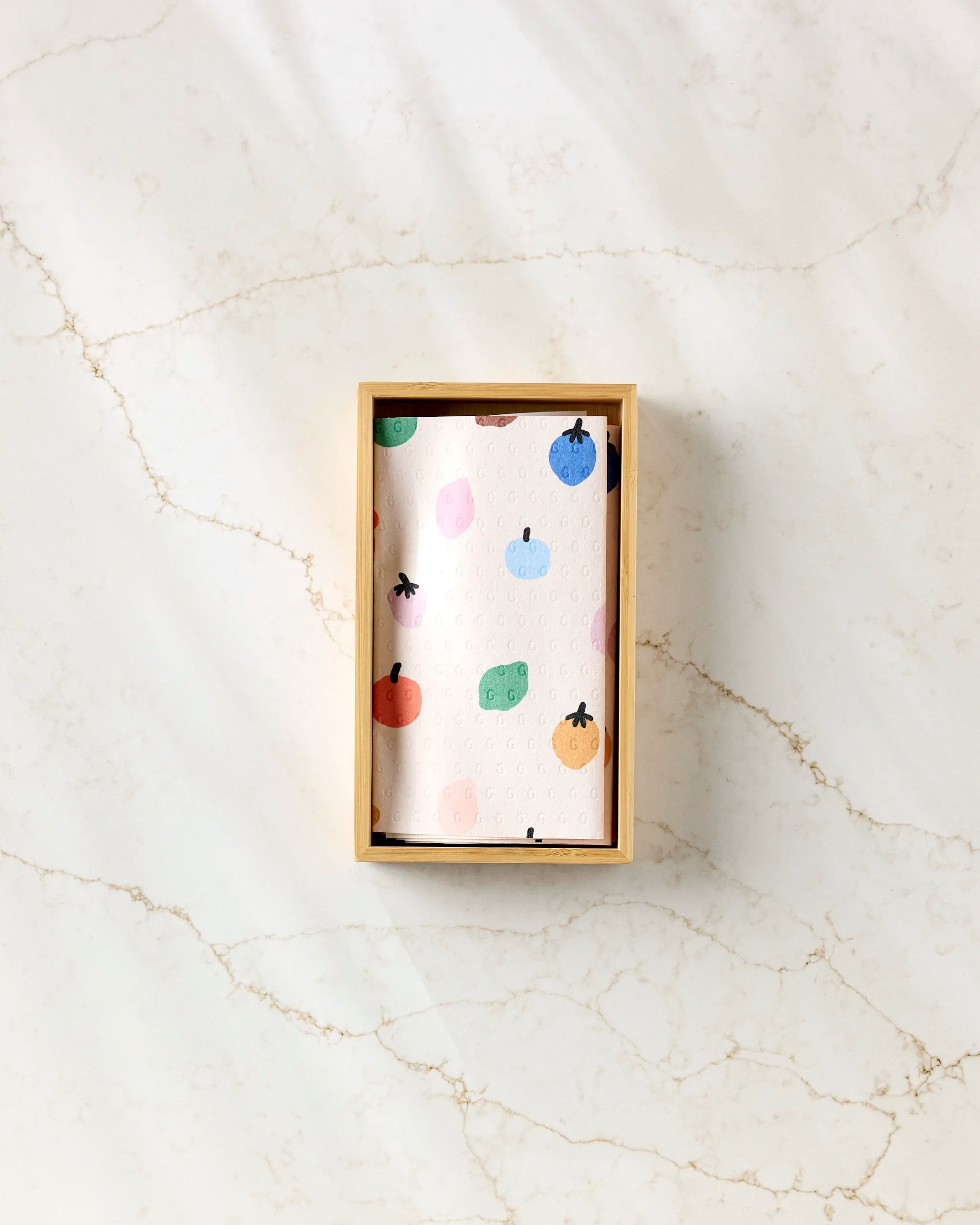 The "Fruit Basket" reusable towel by Jennifer Bouron features colorful fruit and cheese illustrations on a white background—an eco-friendly alternative to paper towels, presented in a rectangular wooden box on a light marble surface.