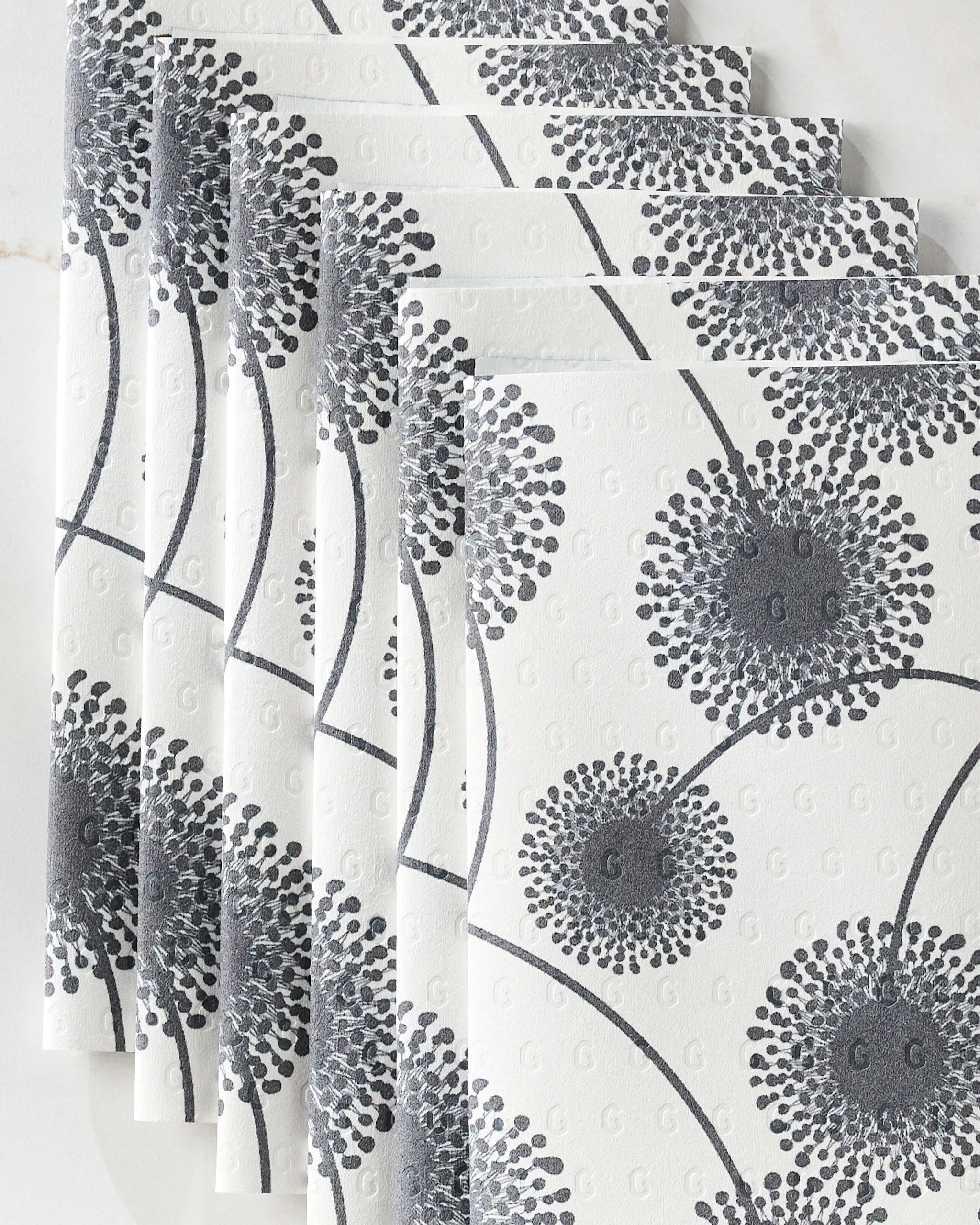 GEOMETRY’s Fully Bloomed white paper napkins feature a raised gray dandelion pattern and curved stems, arranged in a fanned stack—an elegant, eco-friendly touch for any setting.