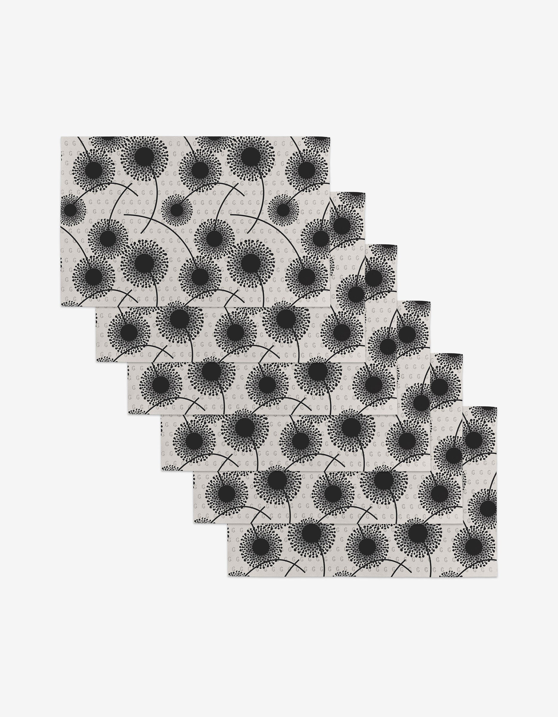 Six Fully Bloomed rectangular reusable placemats, featuring a black dandelion pattern on light gray, are arranged in a cascading diagonal stack on a white surface.