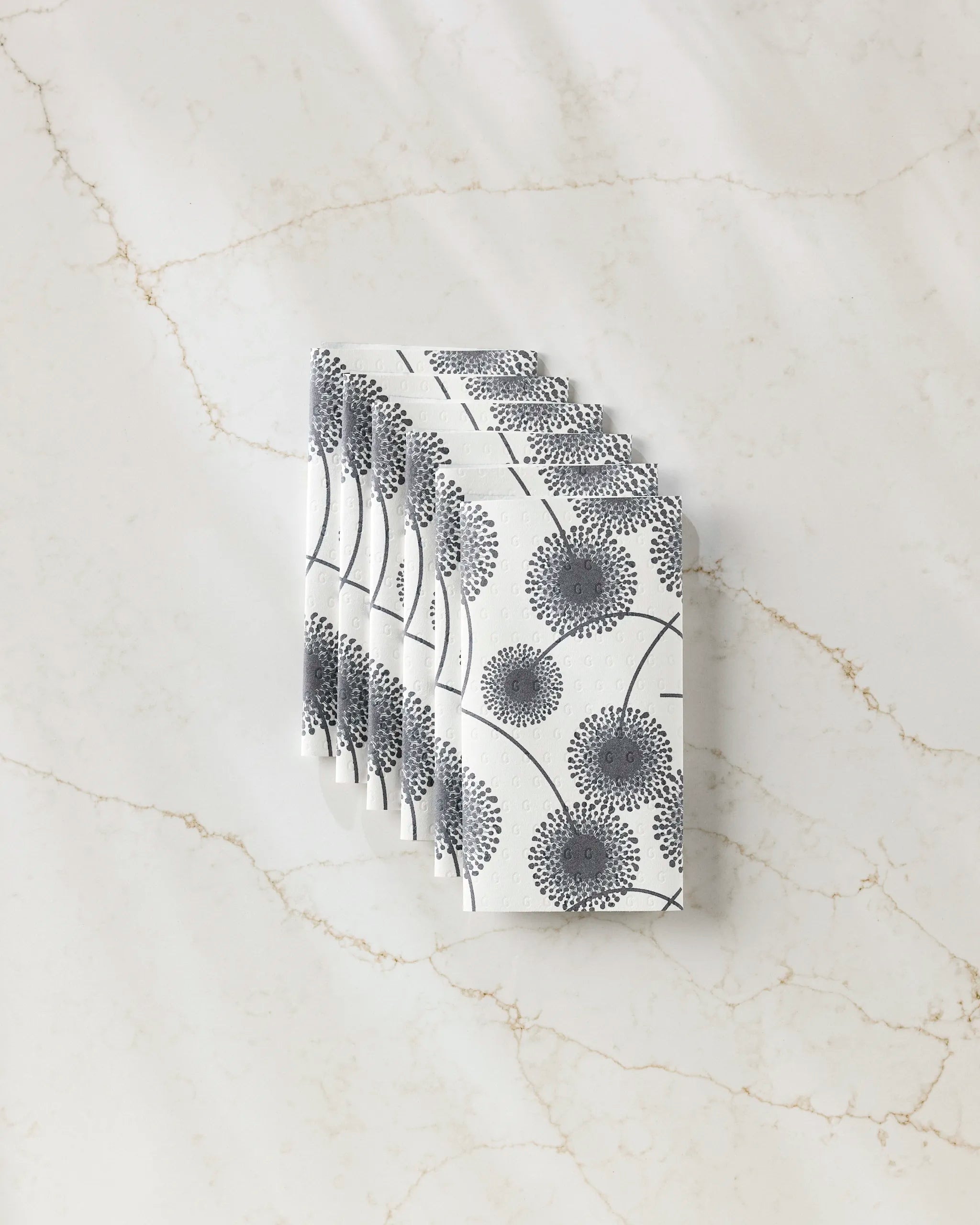 A stack of GEOMETRY’s Fully Bloomed white reusable paper towels, featuring a black dandelion print, is neatly displayed on a white marble surface with light brown veining.