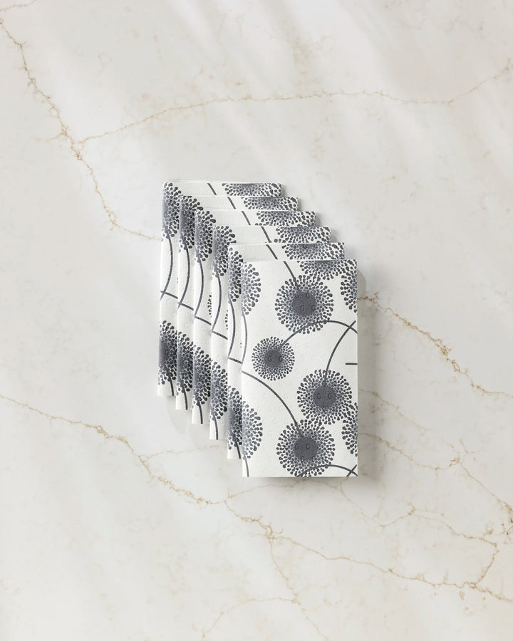 A stack of GEOMETRY’s Fully Bloomed white reusable paper towels, featuring a black dandelion print, is neatly displayed on a white marble surface with light brown veining.