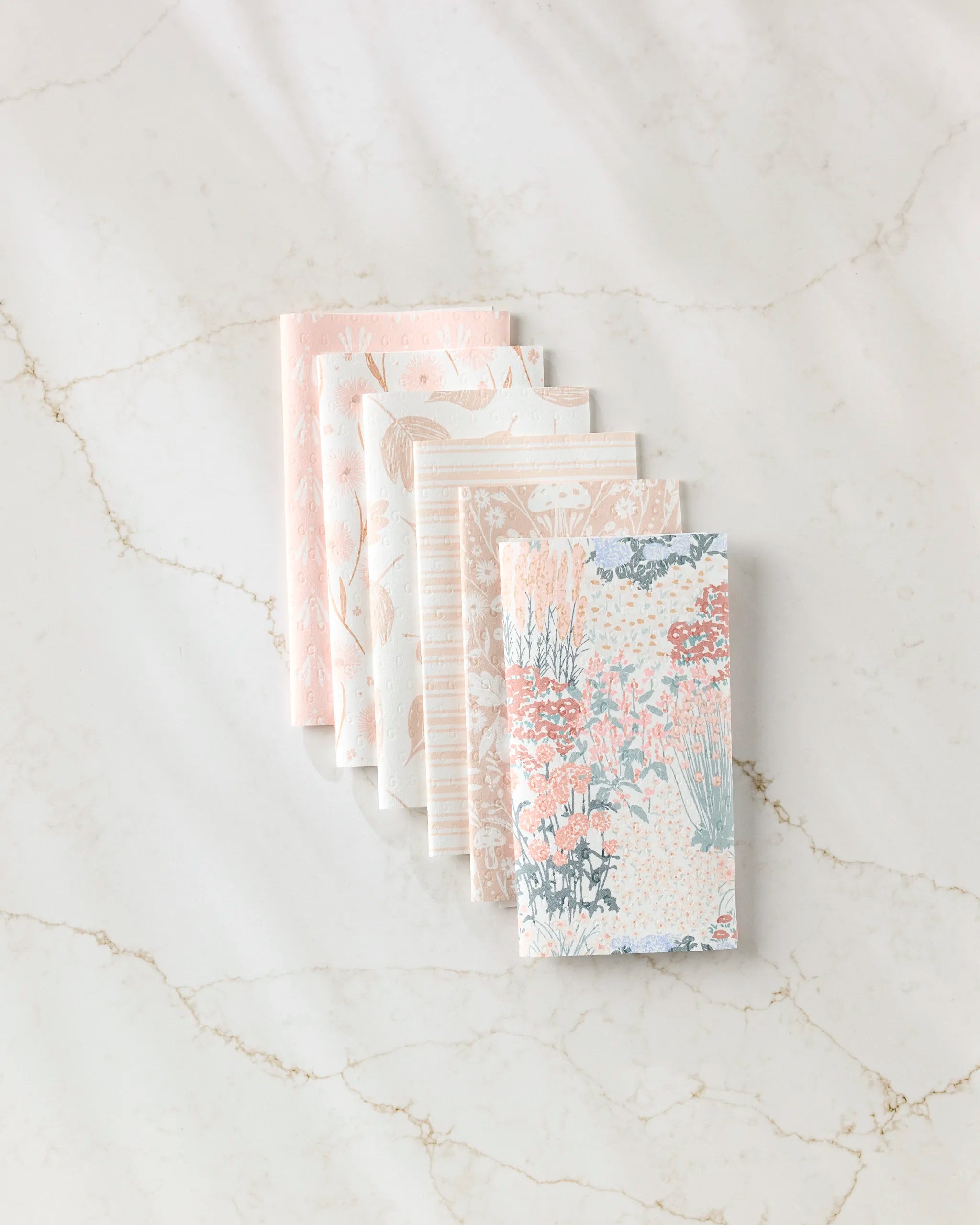 Five folded "Garden Bloom" reusable paper towels by Hufton Studio, featuring delicate floral and geometric patterns in soft pastels, are arranged in a staggered stack on a light marble surface.