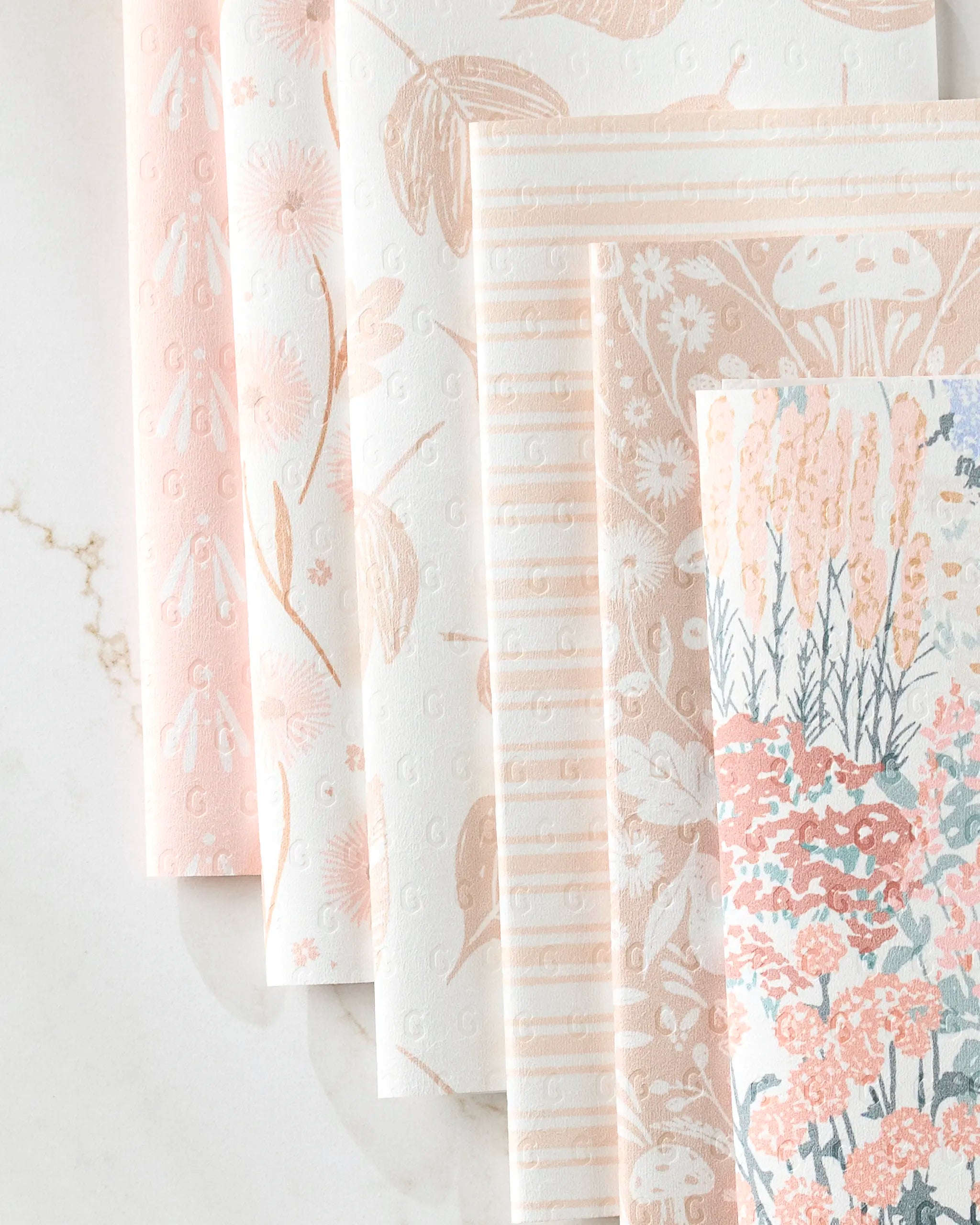 Five sheets of Hufton Studio's Garden Bloom recycled paper towels are fanned out on marble, featuring pastel floral, leaf, stripe, and mushroom patterns in pink, peach, and cream.