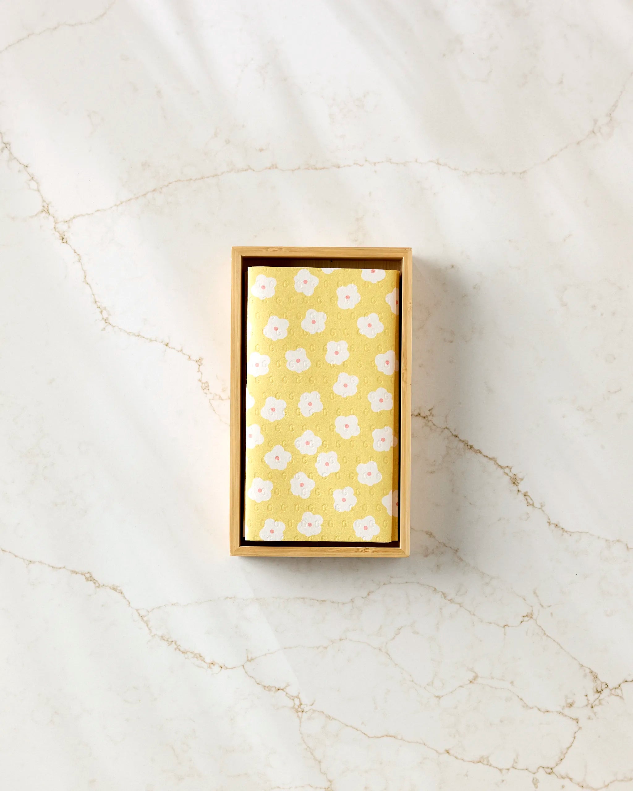 A rectangular Sara Mari wooden box holds the Garden Crush yellow eco-friendly kitchen towel with a white and pink floral pattern, displayed on a light marble surface with subtle veining.