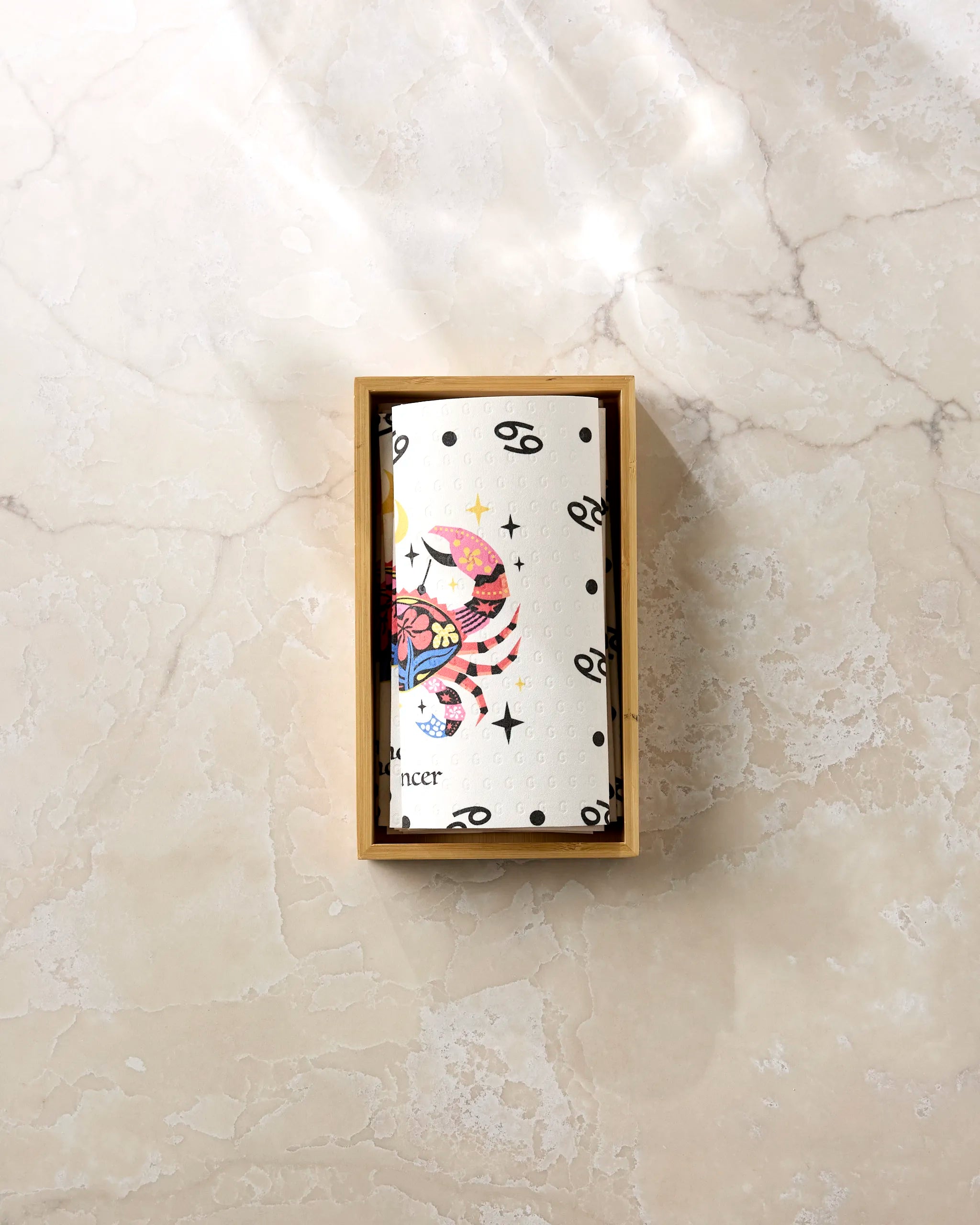 The Gentle Cancer by Heather Zhou features a vibrant Cancer zodiac design with black symbols, housed in a small wooden box—ideal for storing eco-friendly kitchen towels or reusable paper towels on your countertop.