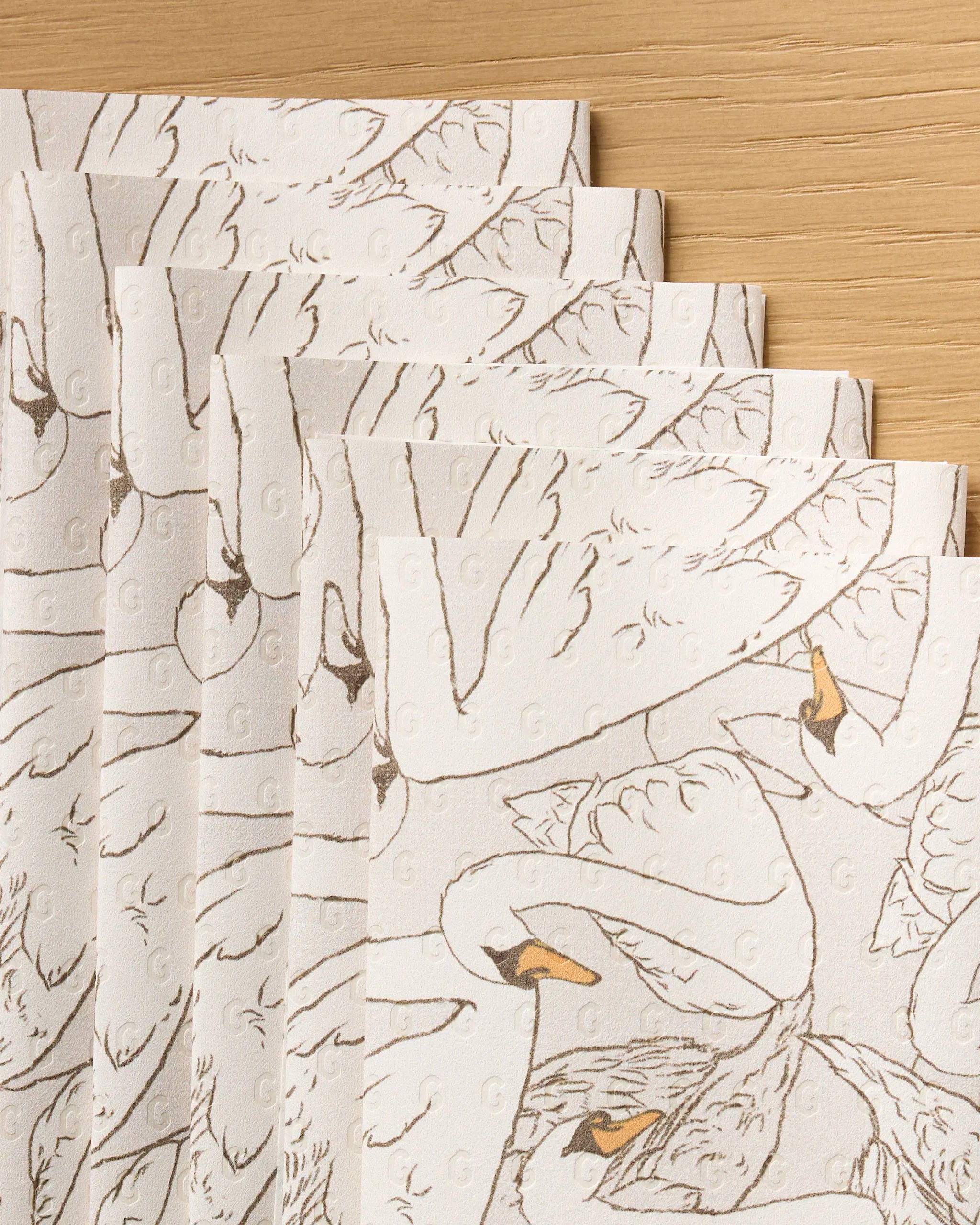 A close-up of Geometry's Gentle Strength recycled paper towels, featuring swan prints and subtle embossed dots, overlapping on a light wooden surface.