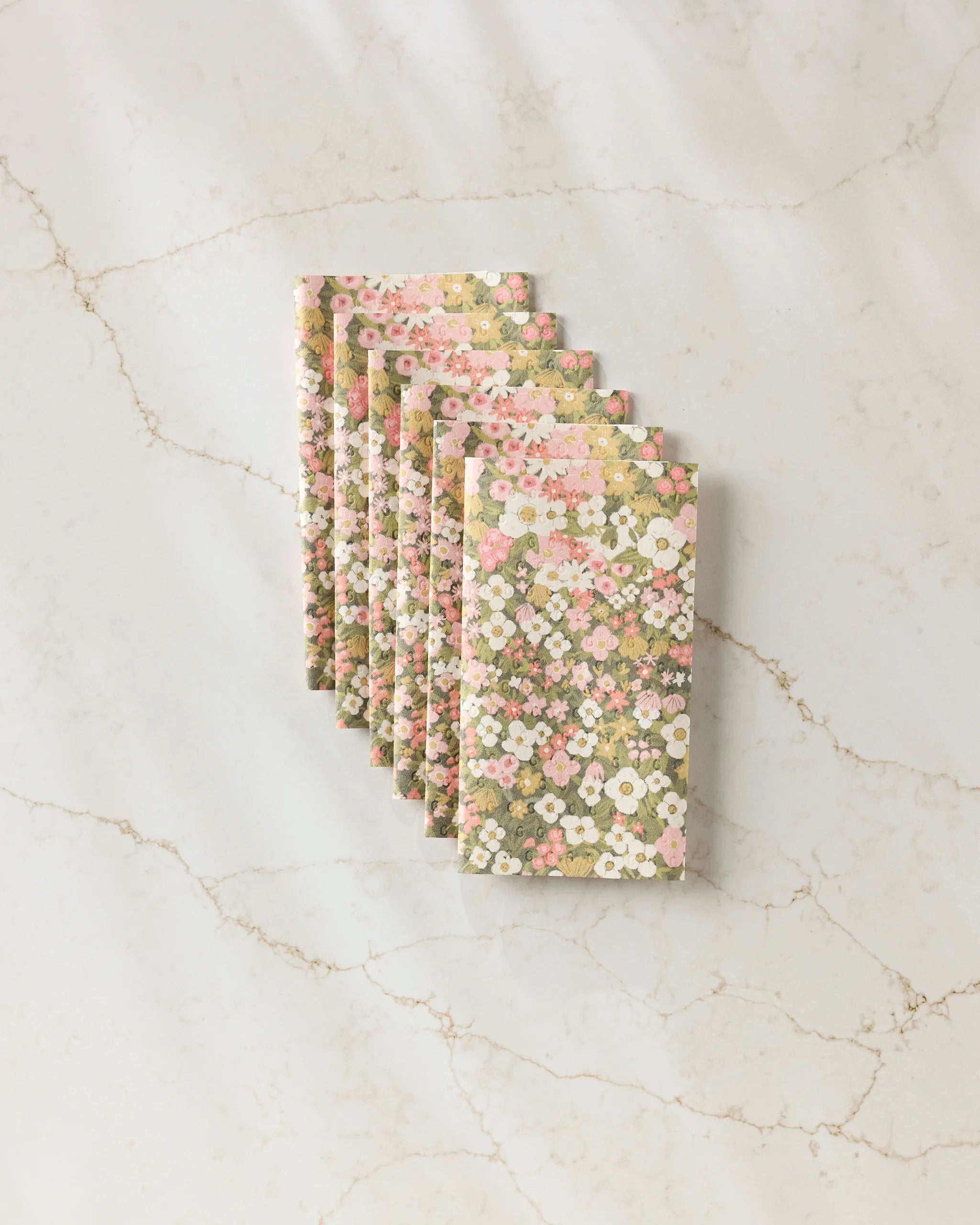 Seven Gilded Garden napkins by Lisa Nelson, featuring pink, white, and green floral patterns, are stacked on a white marble surface. These reusable Not Paper Towels offer an eco-friendly alternative to disposable options.