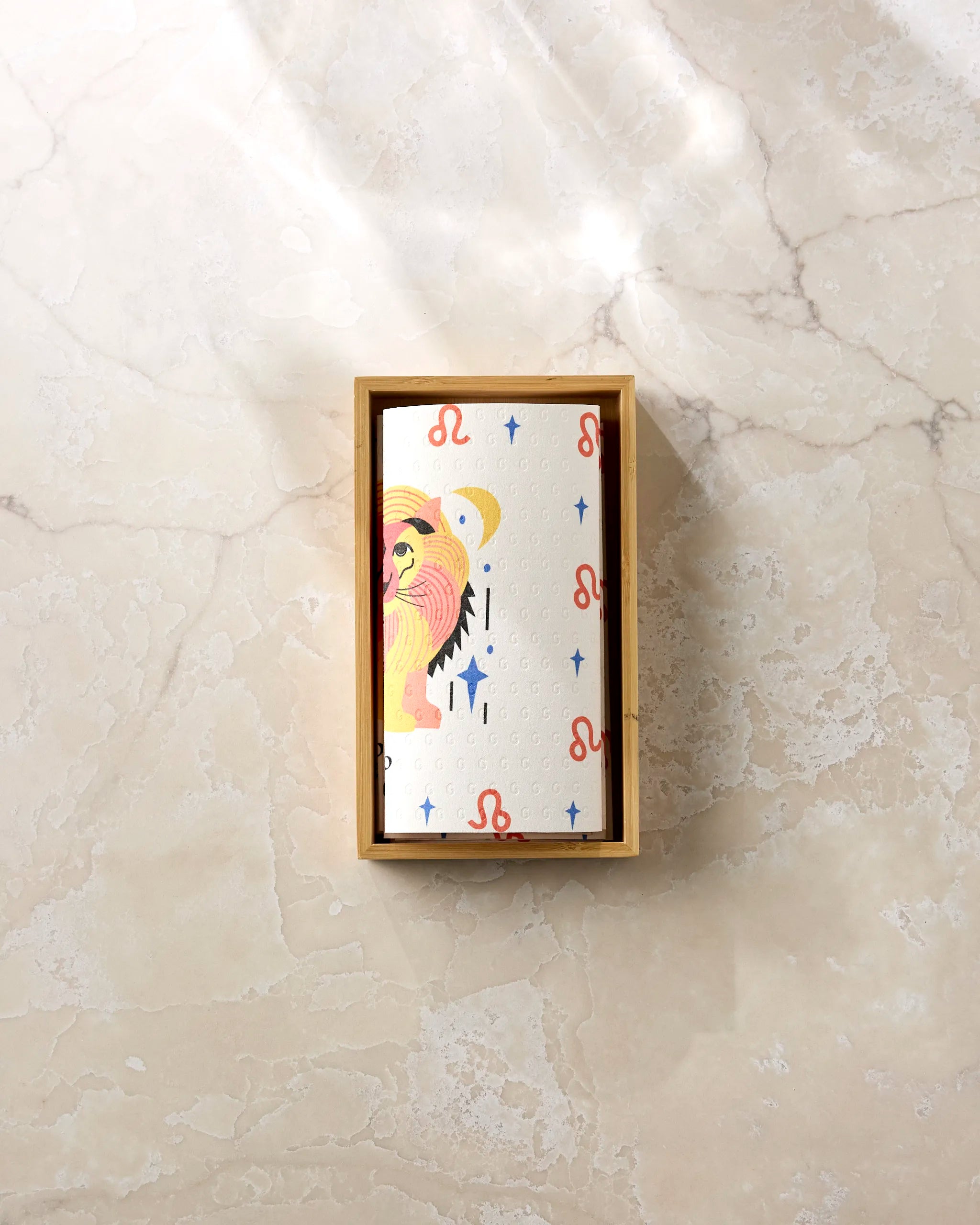 Heather Zhou’s Glowing Leo playing card, with a stylized lion, stars, and red-blue Leo symbols on cream, rests in a rectangular box on marble beside eco-friendly paper towels for a sustainable touch.