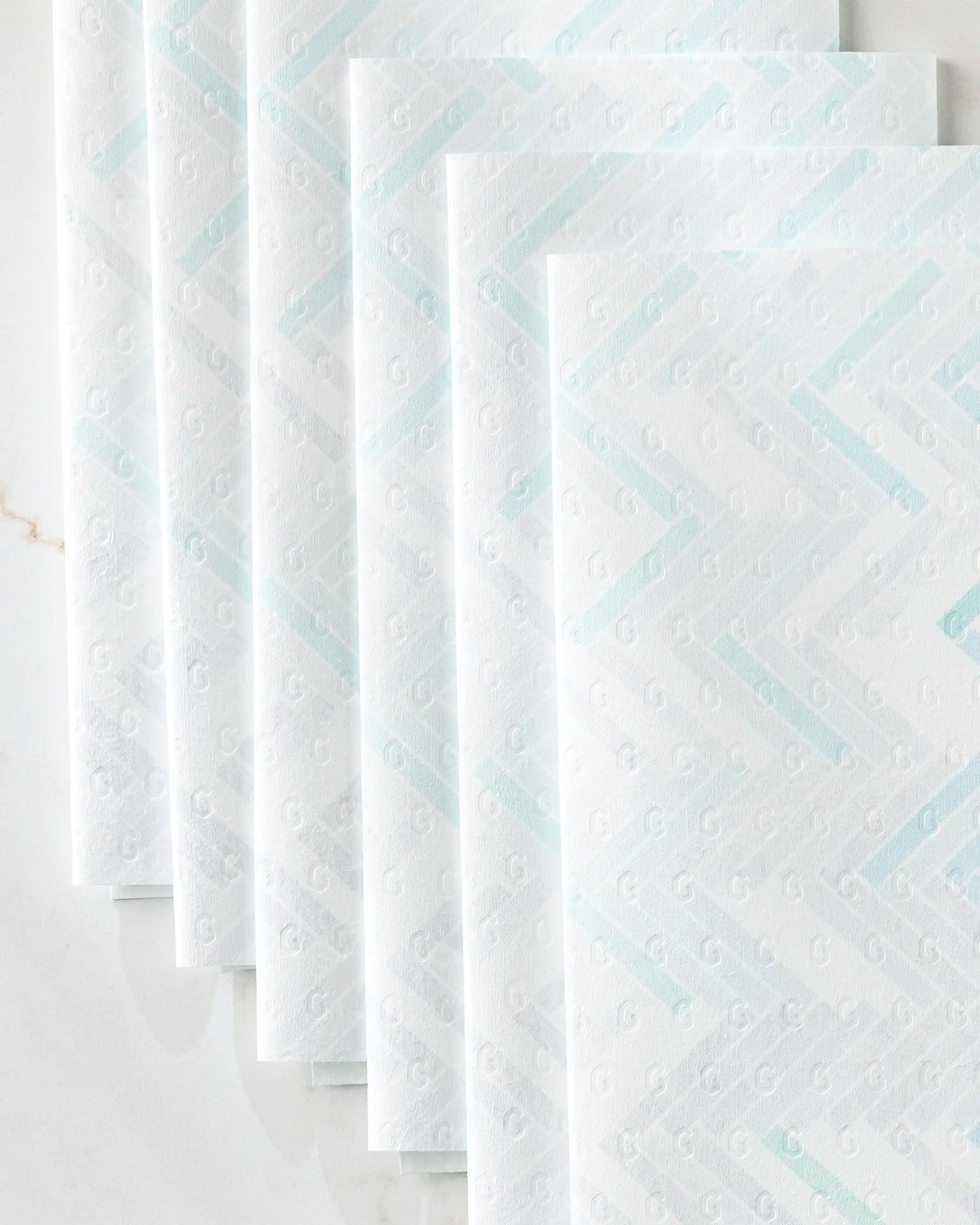 Five “Going Up” reusable paper towels by GEOMETRY, featuring a subtle zigzag and small G designs in pastel blue and gray, are stacked diagonally on a white marble surface.