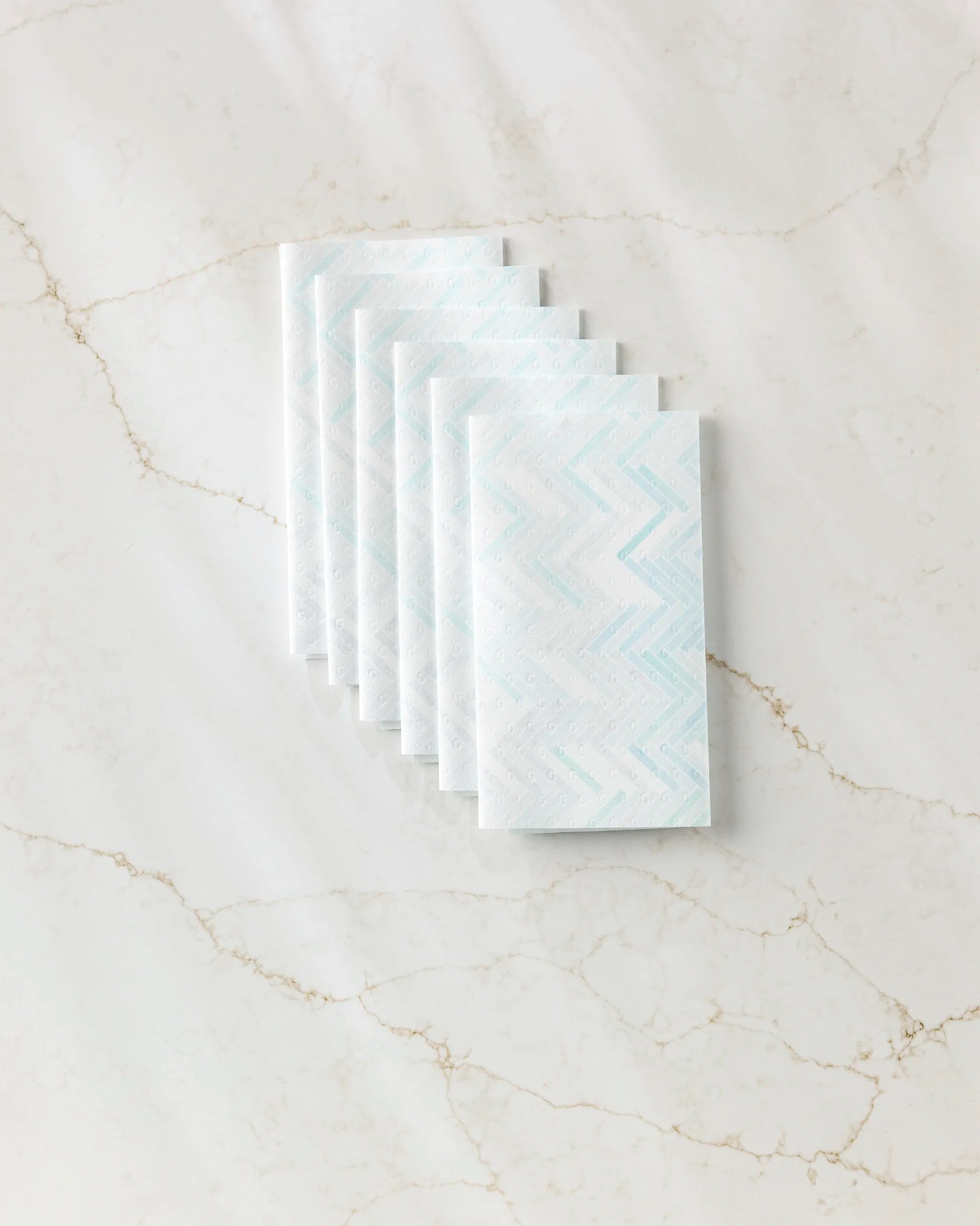 Six folded GEOMETRY "Going Up" reusable paper towels with a blue zigzag pattern are neatly stacked on a white marble surface with subtle brown veins.
