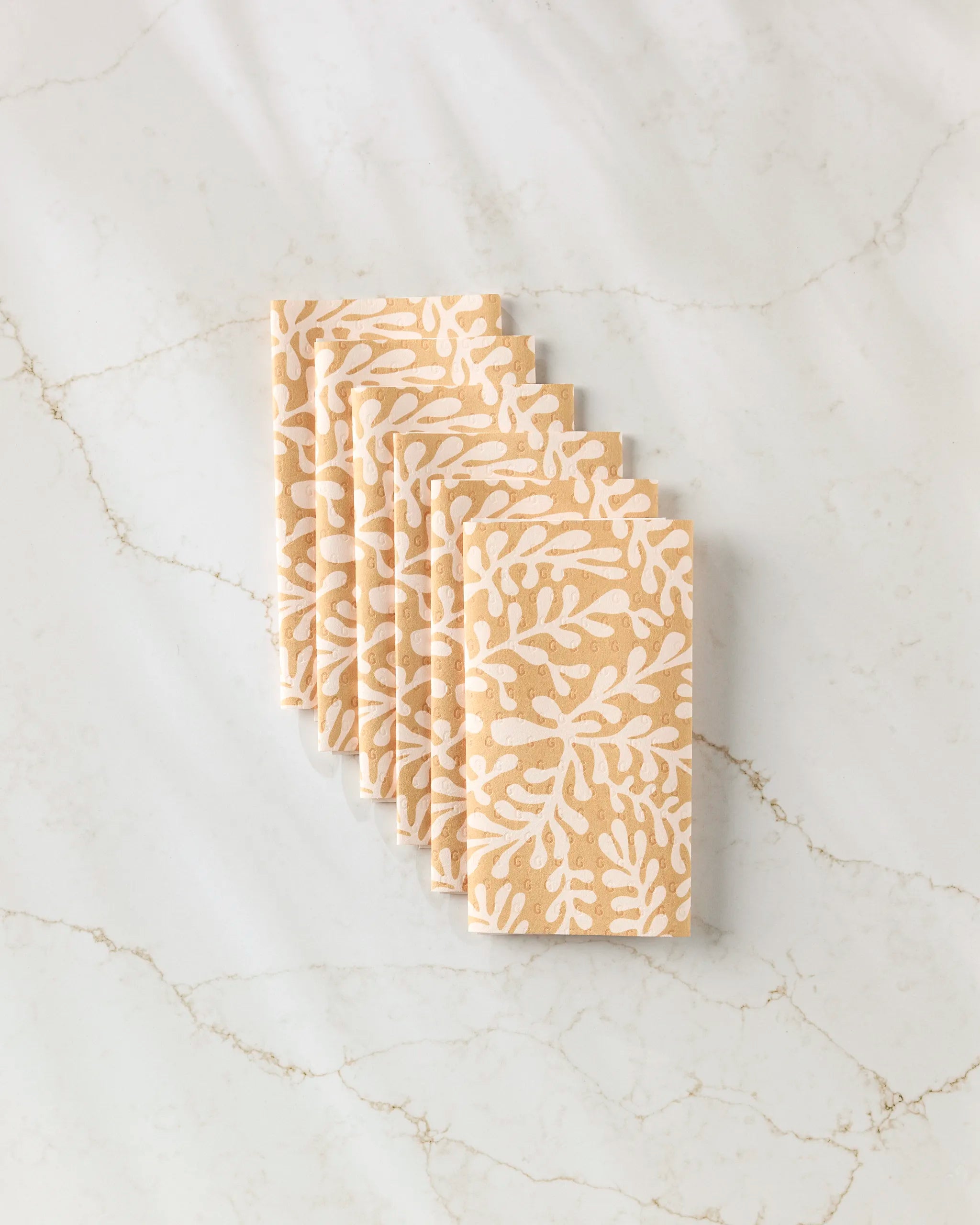 Six GEOMETRY “Golden Fall” napkins, beige with a white leaf pattern and resembling reusable paper towels, are stacked in a fan arrangement on a light marble surface with subtle gray and tan veining.
