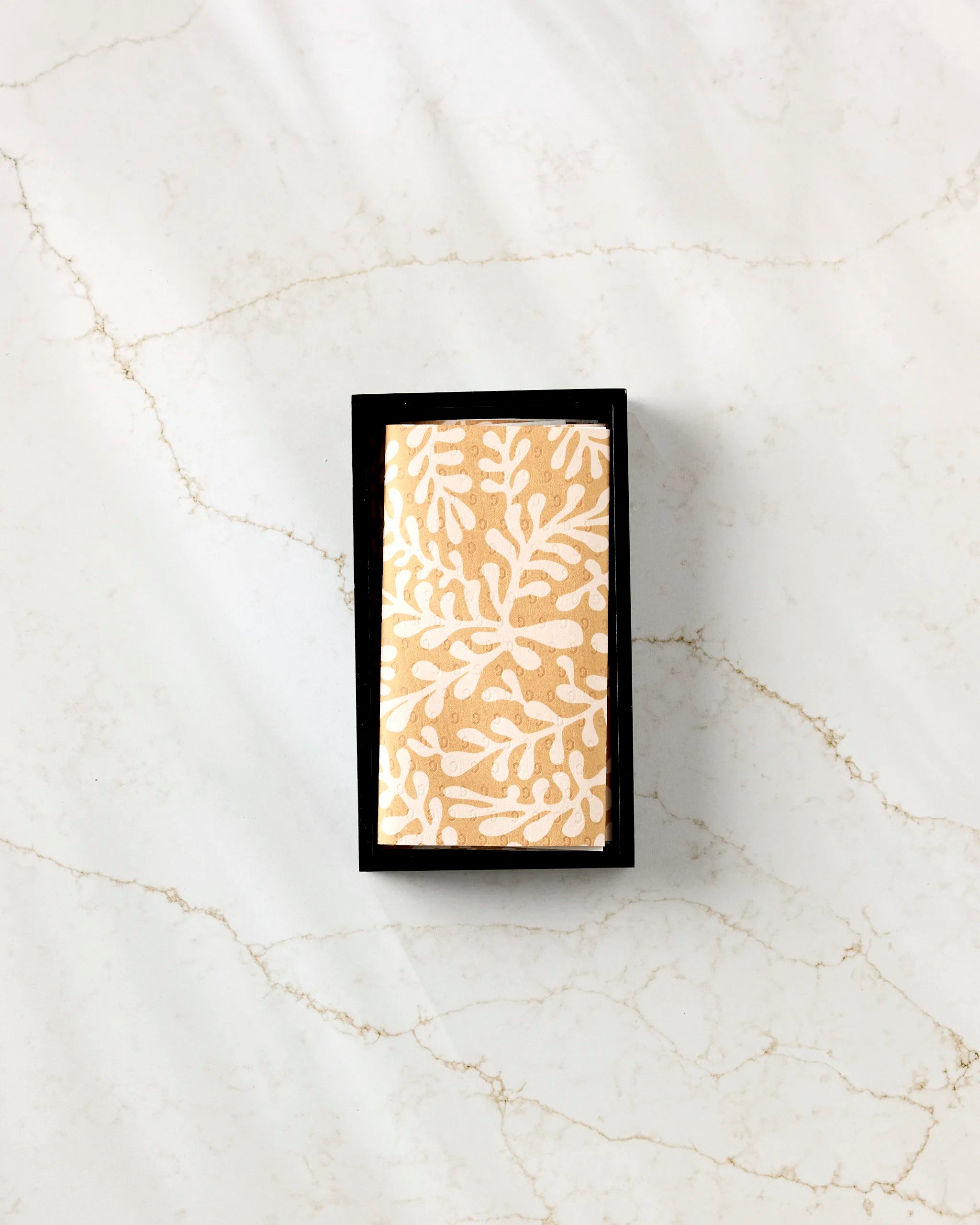 The GEOMETRY Golden Fall rectangular bar soap, featuring a white leaf design, rests in a black dish on beige-veined white marble beside a stack of eco-friendly paper towels.