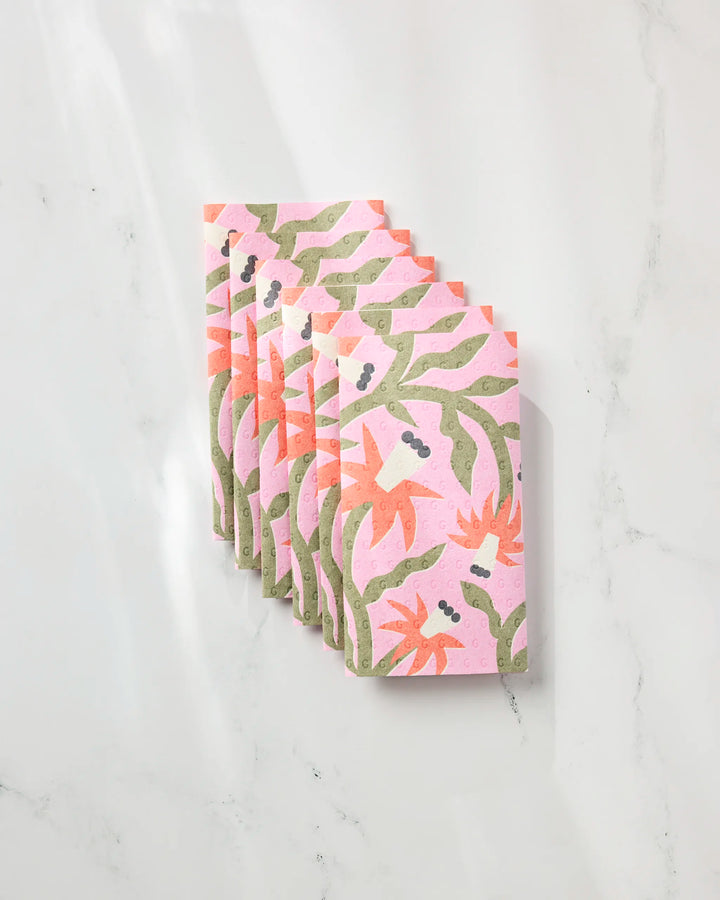 Six folded Claire Iglesias "Graphic Flowers" reusable paper towels, featuring green and orange floral patterns on a pink background, are neatly arranged in a row on a white marble surface.
