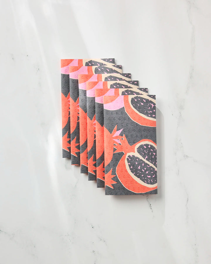 Eight rectangular Graphic Pomegranates cards by Claire Iglesias, showcasing stylized pomegranates in pink, red, and gray hues, are fanned out on a white marble surface, evoking the look of eco-friendly kitchen towels.