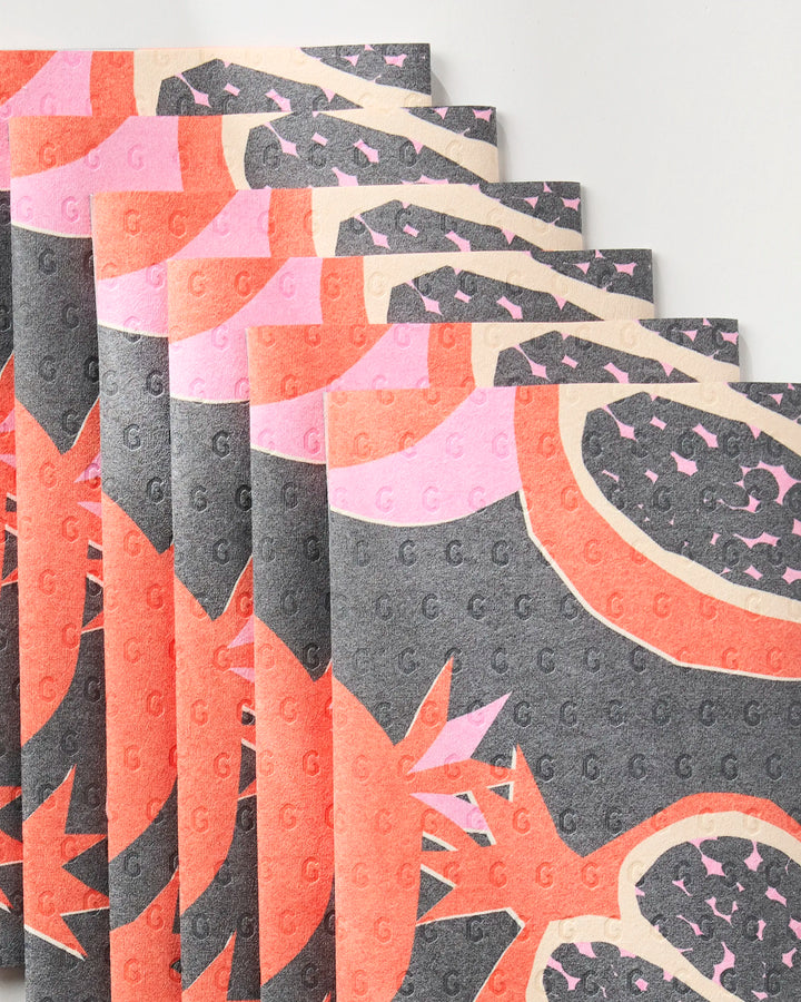 The "Graphic Pomegranates" kitchen towels by Claire Iglesias feature eco-friendly, textured fabric with bold red, pink, and black abstract pomegranate patterns and repeated “G” imprints on gray.