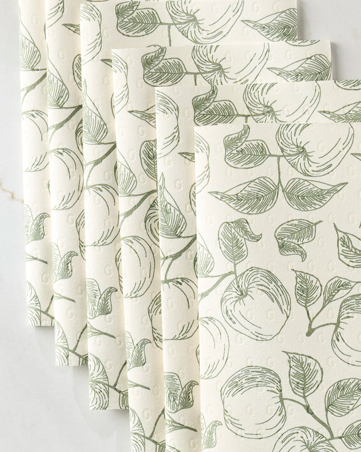 Five cream-colored Gravenstein Apple reusable paper towels by Ash Cascade are arranged in a fan shape, each showing a hand-drawn green apple and leaf pattern on a white marble background.