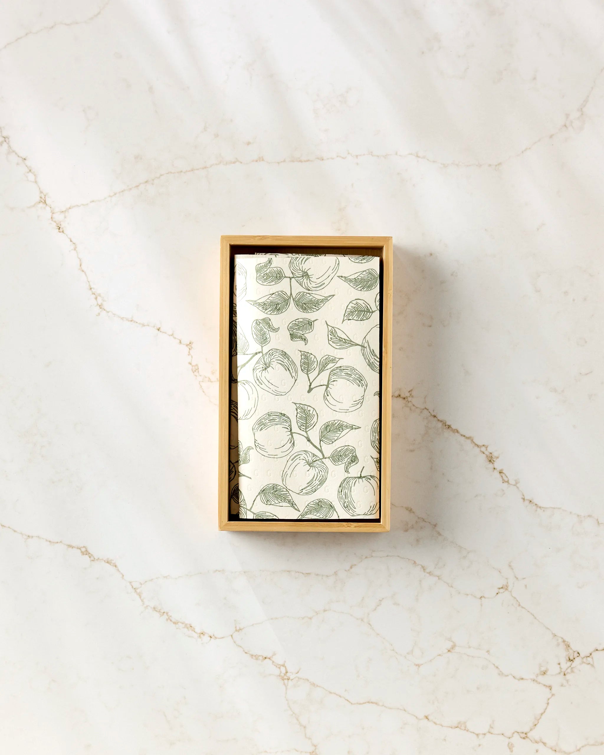 A rectangular Gravenstein Apple box by Ash Cascade, featuring a green fruit and leaf pattern, designed for storing reusable paper towels, sits on a white marble surface with light brown veins.