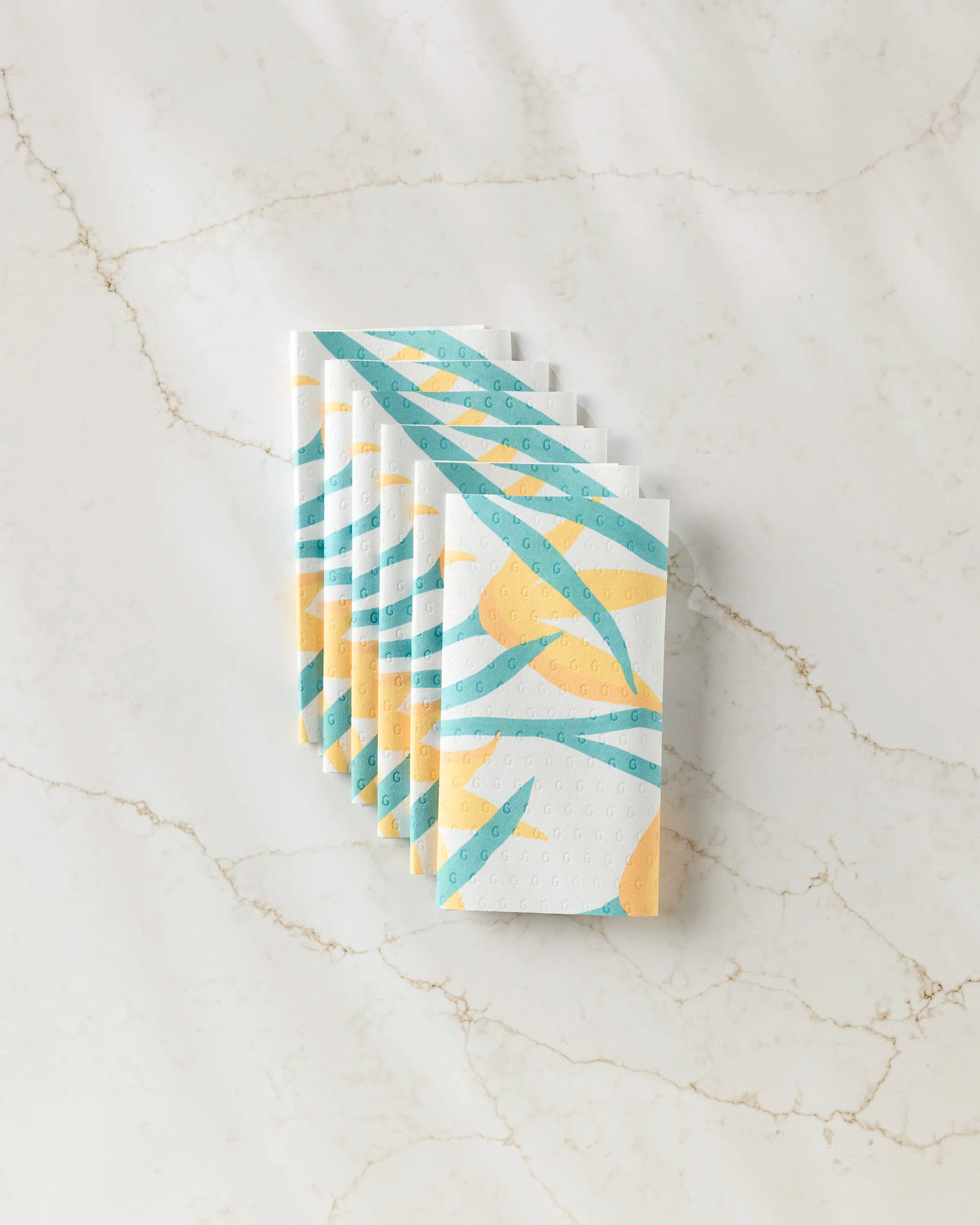 Six folded, eco-friendly GEOMETRY Growth paper towels with blue and yellow abstract patterns are neatly arranged in a row on a white marble surface with subtle brown veining.