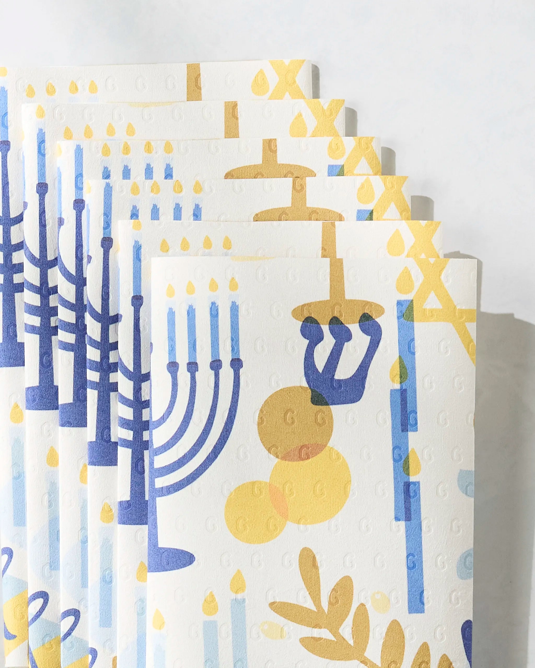 Hanukkah is Here by Geometry: Festive, eco-friendly paper towels adorned with blue menorahs, yellow dreidels, gold coins, candles, and olive branches—perfect for celebrating the Hanukkah holiday.