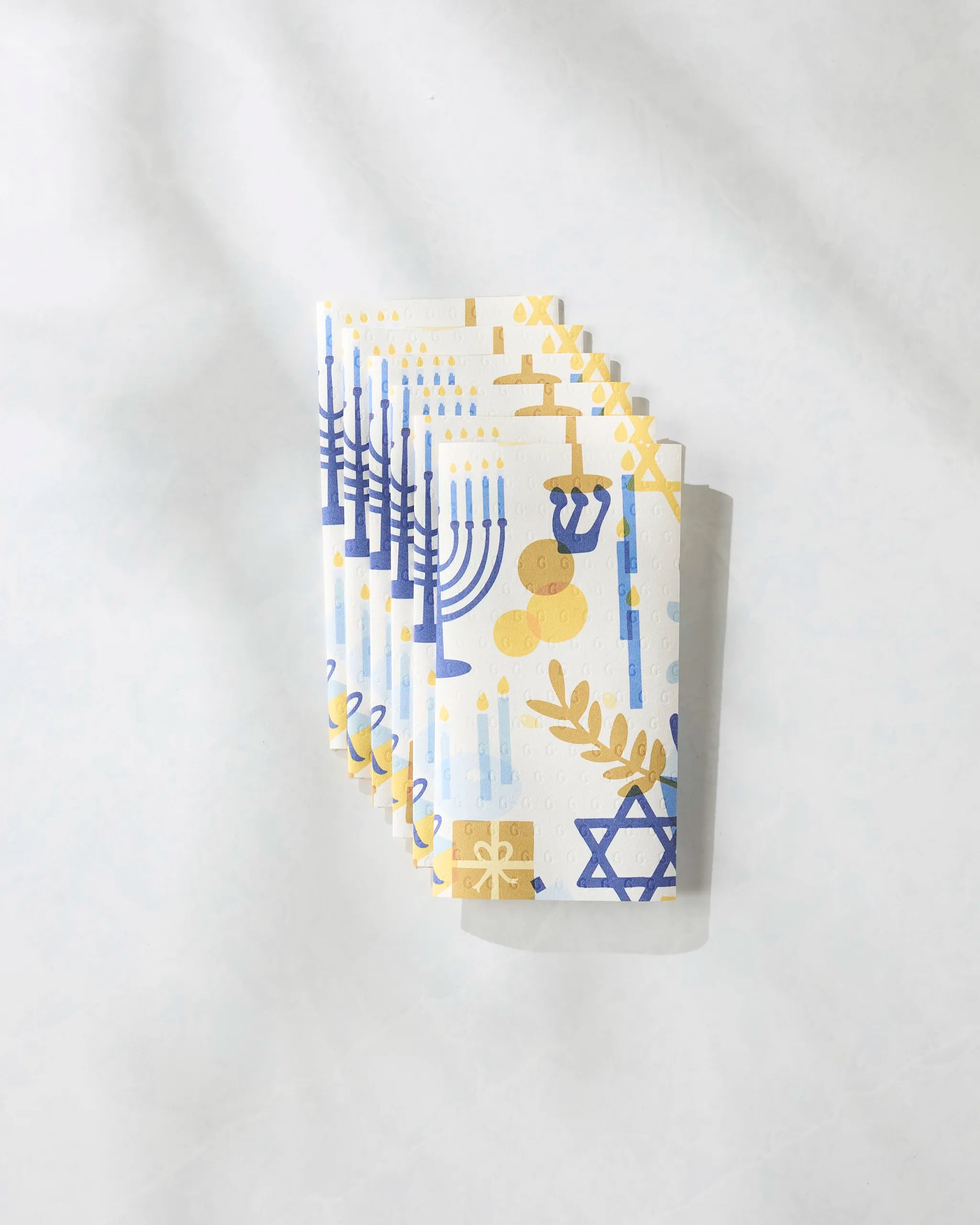 Four "Hanukkah is Here" napkins by Geometry, made from eco-friendly paper towels, display blue and yellow menorahs, dreidels, candles, gift boxes, and Stars of David, fanned out on a light surface.