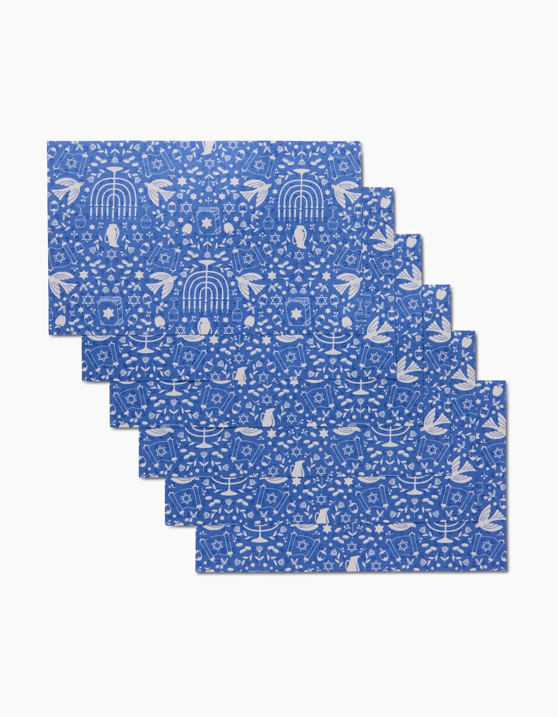 Six blue Hanukkah Sameach placemats by Megan Kampa feature white folk-art birds, leaves, and flowers. Eco-friendly, reusable, and machine washable, theyre shown overlapping on a white background.