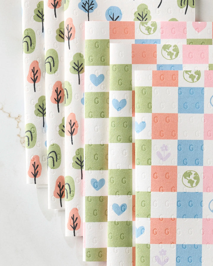 Kayla Ann’s Happy Checkers set includes four eco-friendly kitchen towels: one with pastel blue-green trees, and three with checkered hearts, flowers, and globes in pink, purple, blue, and green on a white background.