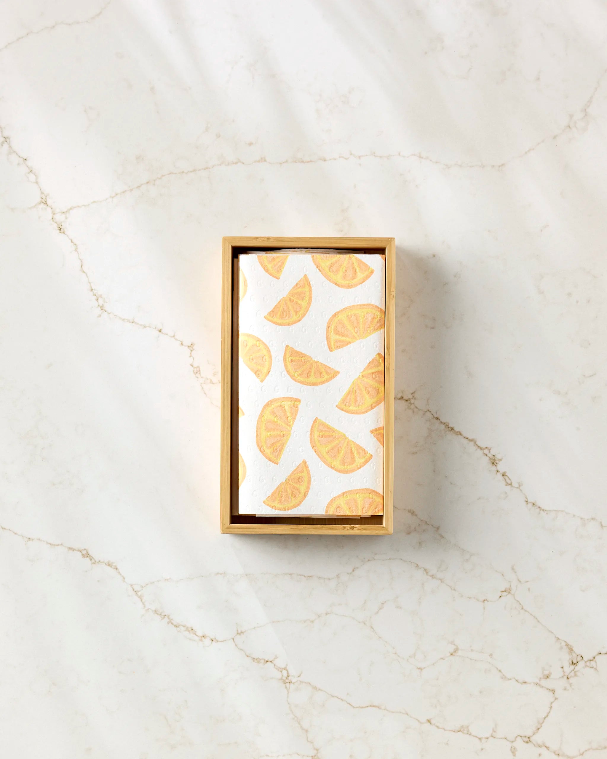 Aubrey Fairchild’s "Here Comes The Sun" eco-friendly kitchen towels, packaged in a rectangular box with a white card and orange citrus slice pattern, rest on subtly veined light marble.