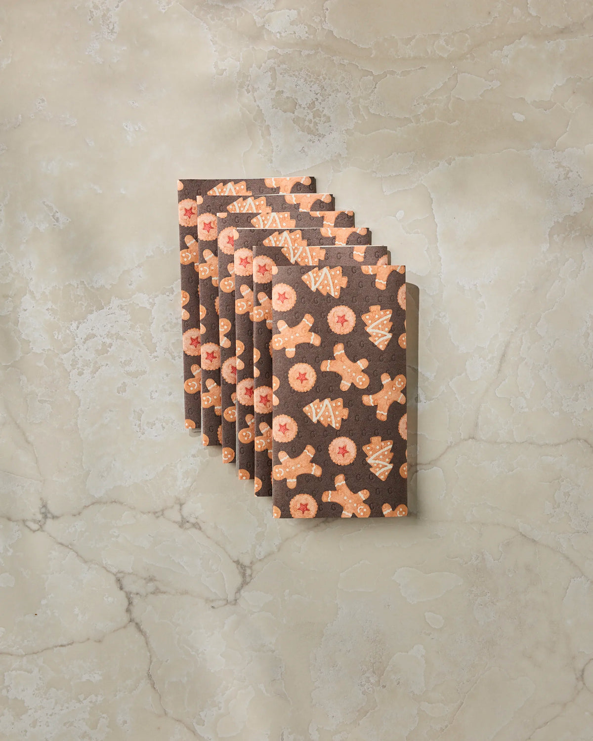 Seven “Holiday Cookies” cards by Geometry, featuring festive cookie patterns on brown post-consumer recycled cardstock, are fanned out on a beige marble surface—offering an eco-friendly alternative to traditional cards.