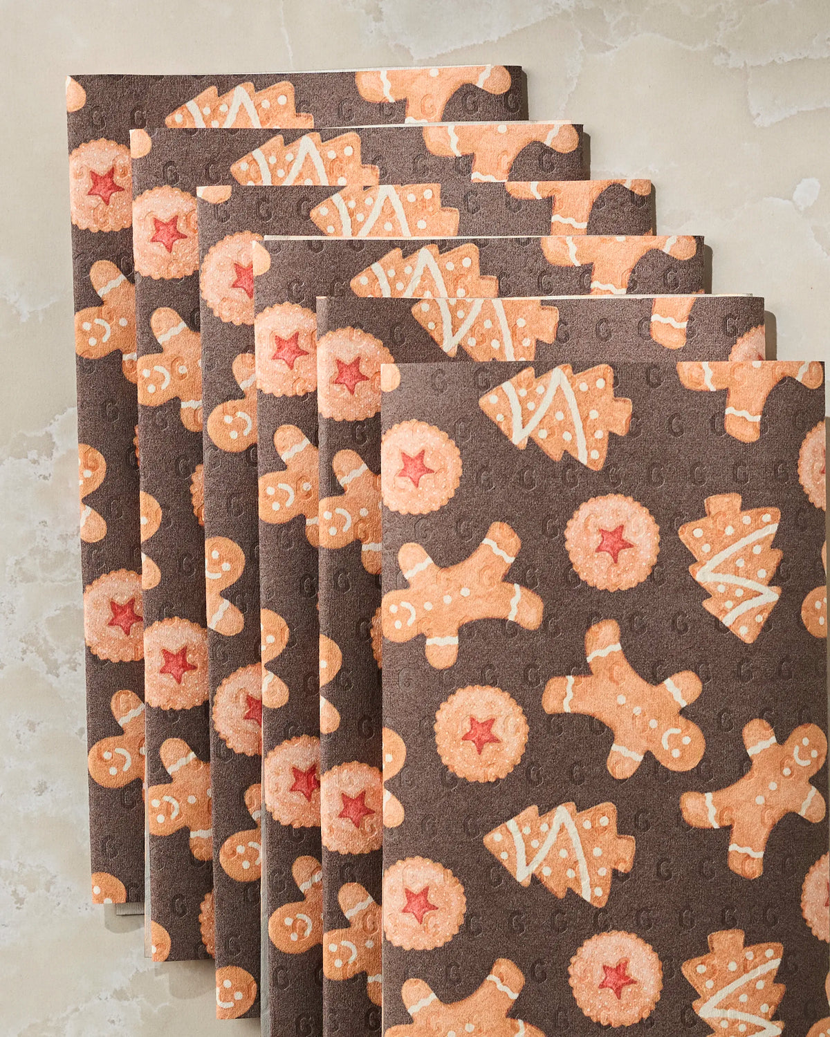 Six brown “Holiday Cookies” reusable paper towels by Geometry, featuring gingerbread men, star-shaped cookies, and round cookies with red centers, are neatly stacked on a light marble surface—an eco-friendly choice made from post-consumer recycled materials.