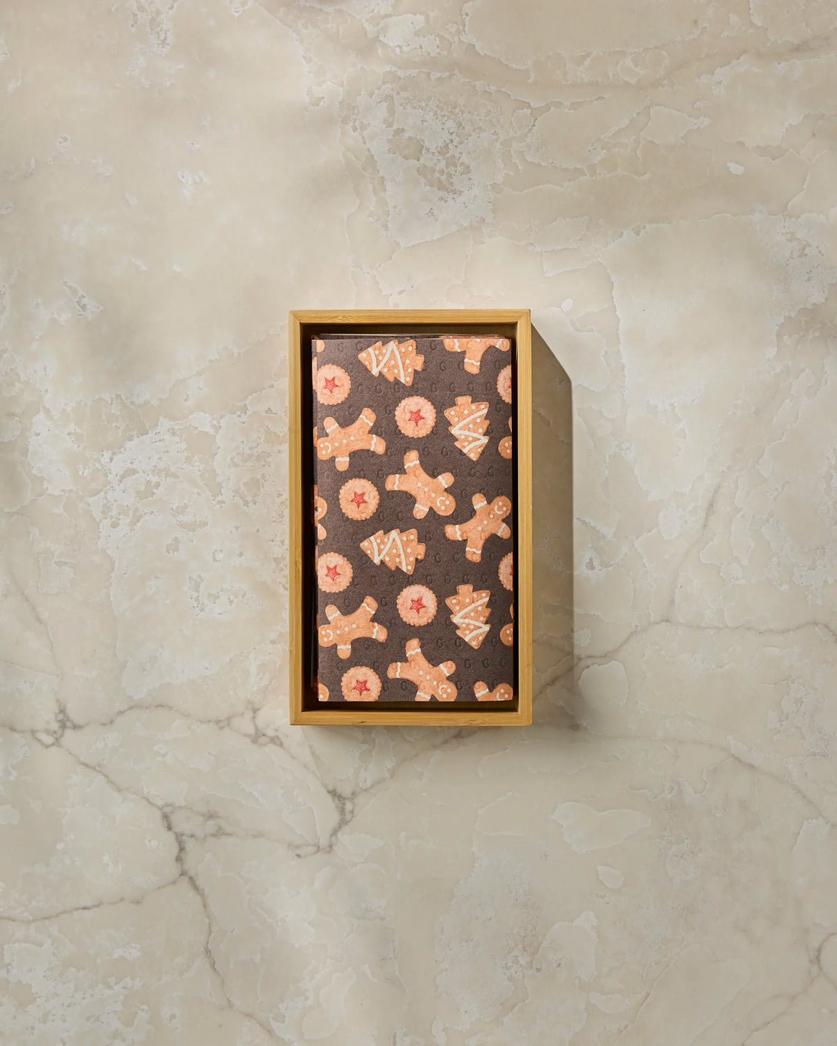 A rectangular wooden box holds “Holiday Cookies” reusable paper towels by Geometry, featuring a festive pattern of gingerbread men, Christmas trees, and stars on a brown background, displayed atop a marbled beige surface. Designed as an environmentally friendly alternative to disposables, this sustainable product is perfect for those seeking eco-conscious kitchen solutions.