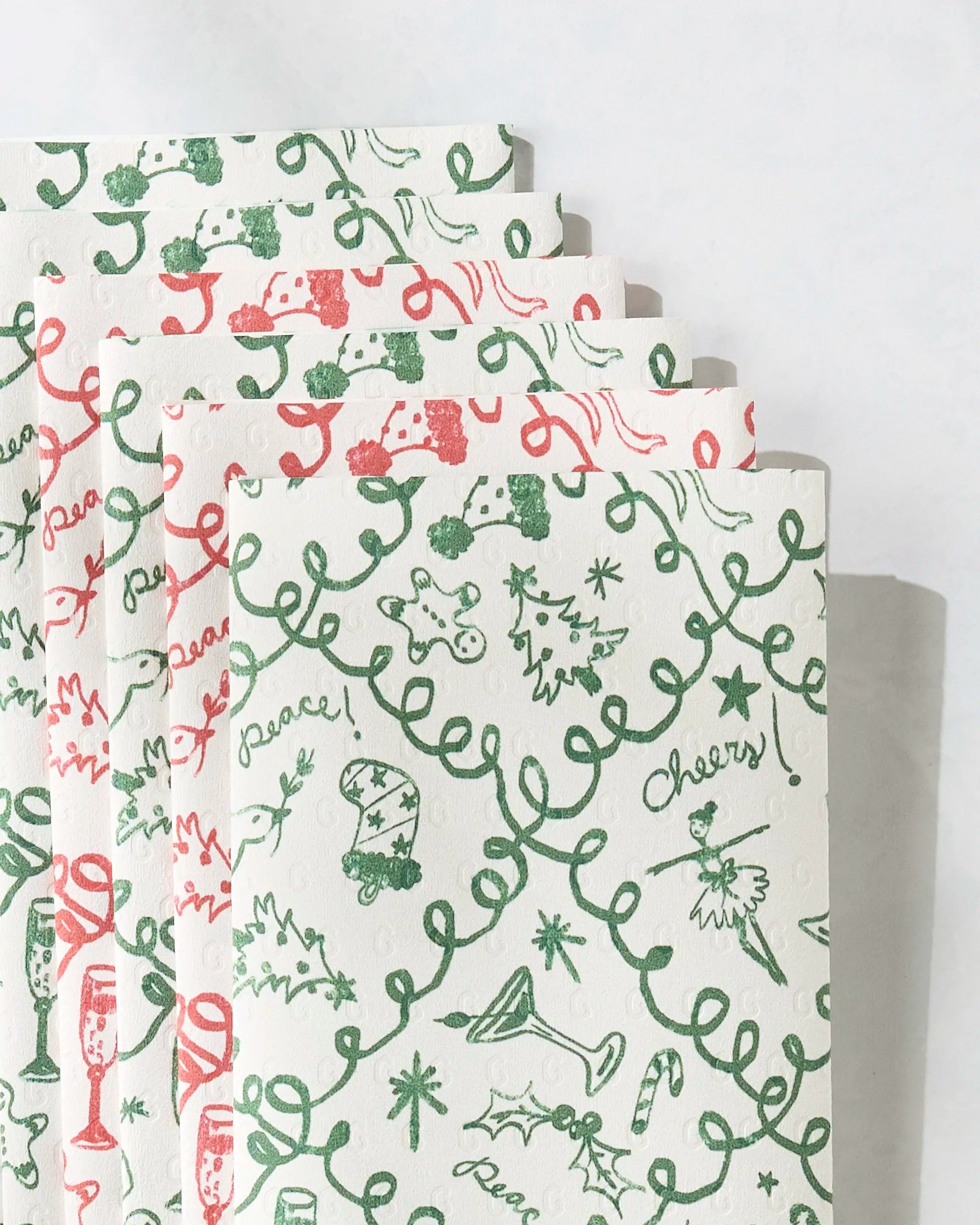 Holiday Doodling by Geometry: five eco-friendly paper napkins featuring cheerful red and green holiday designs—Christmas trees, candy canes, champagne glasses, holly, and festive words—fanned out for a joyous seasonal touch.