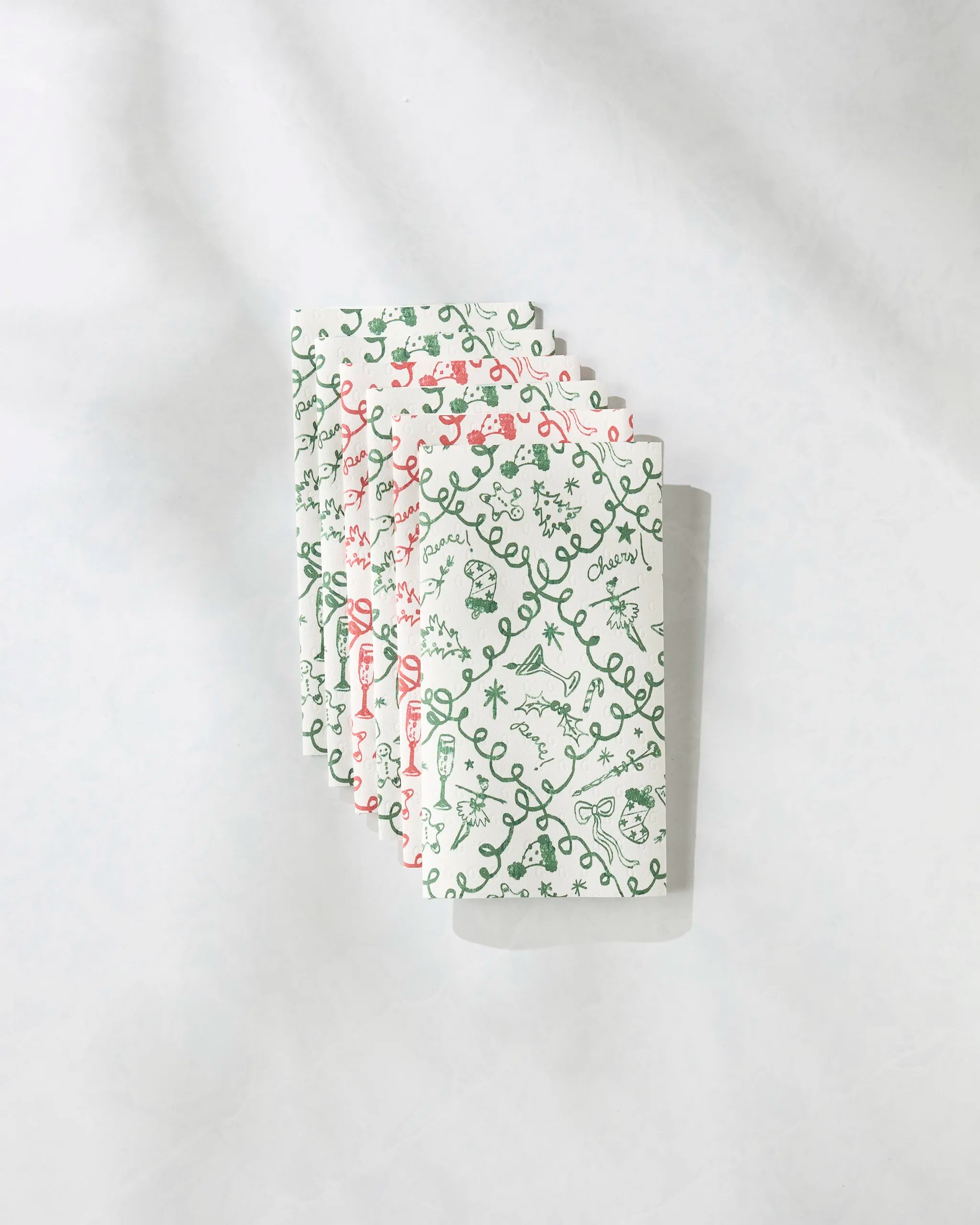 Four "Holiday Doodling" reusable white paper towels by Geometry, featuring playful green and red designs, are arranged in a fanned stack on a light, textured surface.