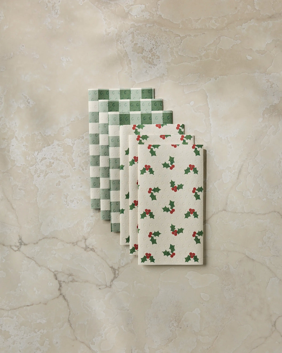 Four folded Geometry Holiday Holly cloth napkins—two featuring a green and white checkerboard pattern and two adorned with a cream background and holly berry print—are arranged on a beige marble surface, offering an eco-friendly alternative to disposable options like reusable paper towels.