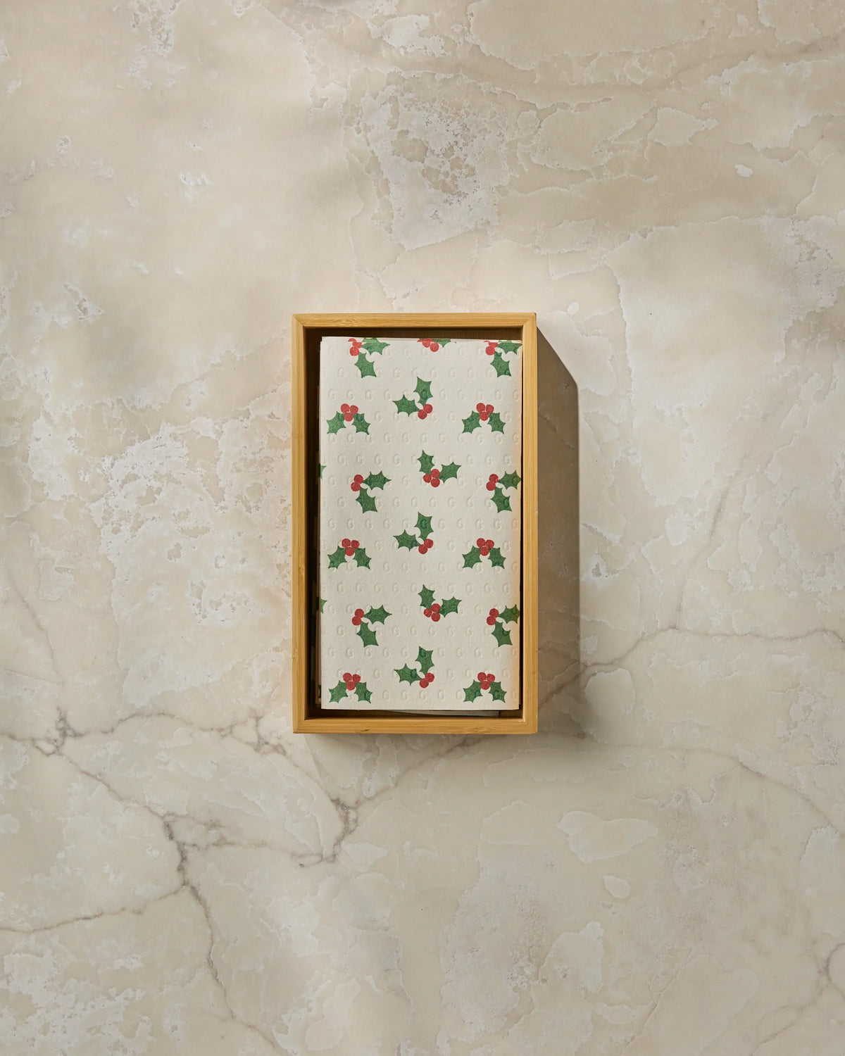 A rectangular wooden box holds Geometry’s Holiday Holly reusable paper towel, featuring a holly leaf and red berry design, displayed on a beige marble surface.
