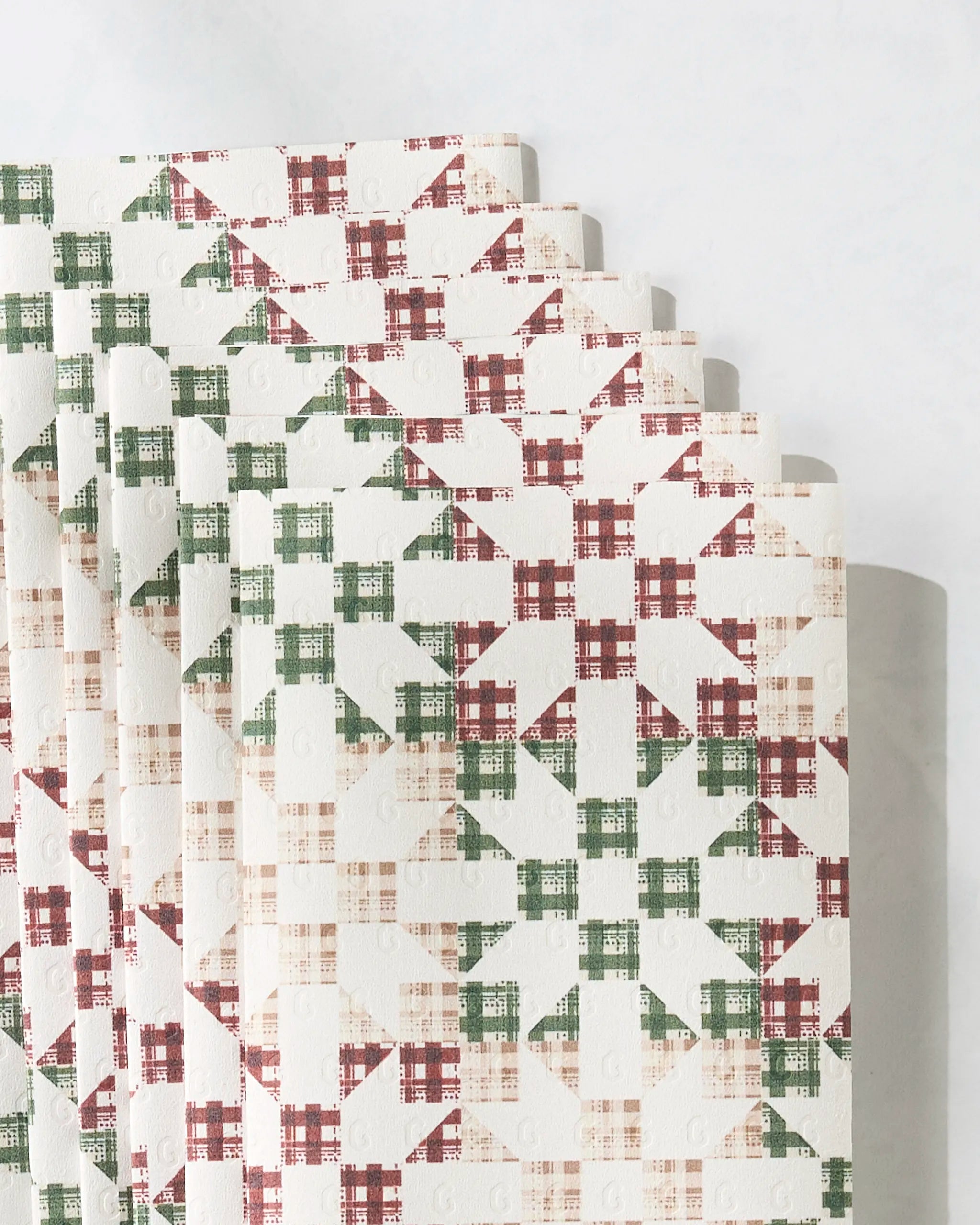 A stack of Geometry’s Holiday Quilt eco-friendly paper towels, showcasing a red, green, and beige plaid pattern, is arranged in a fan shape on a light surface.