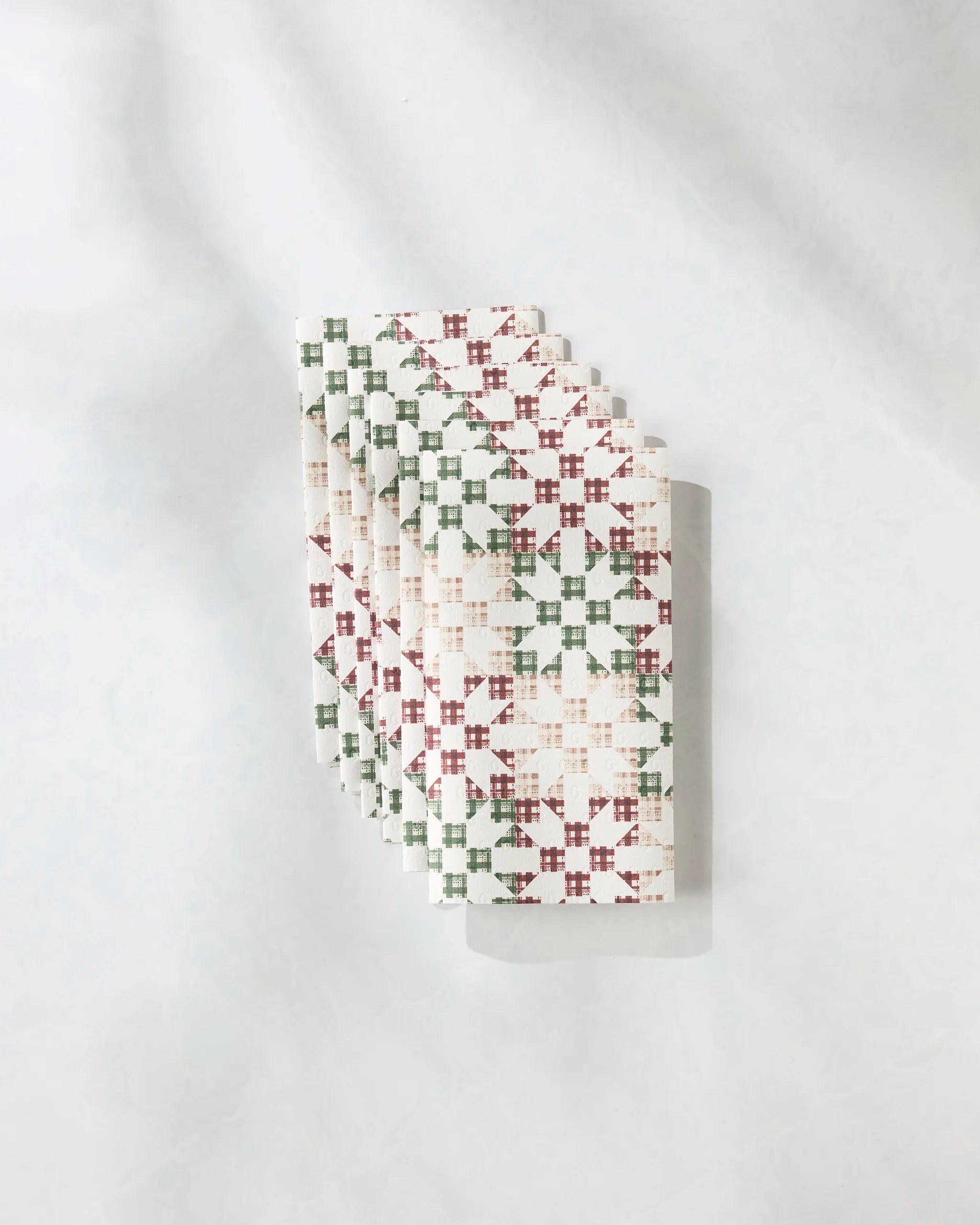 A stack of Geometry Holiday Quilt reusable white paper towels features a red and green geometric cross-stitch pattern, neatly arranged on a light, textured surface with soft shadows.